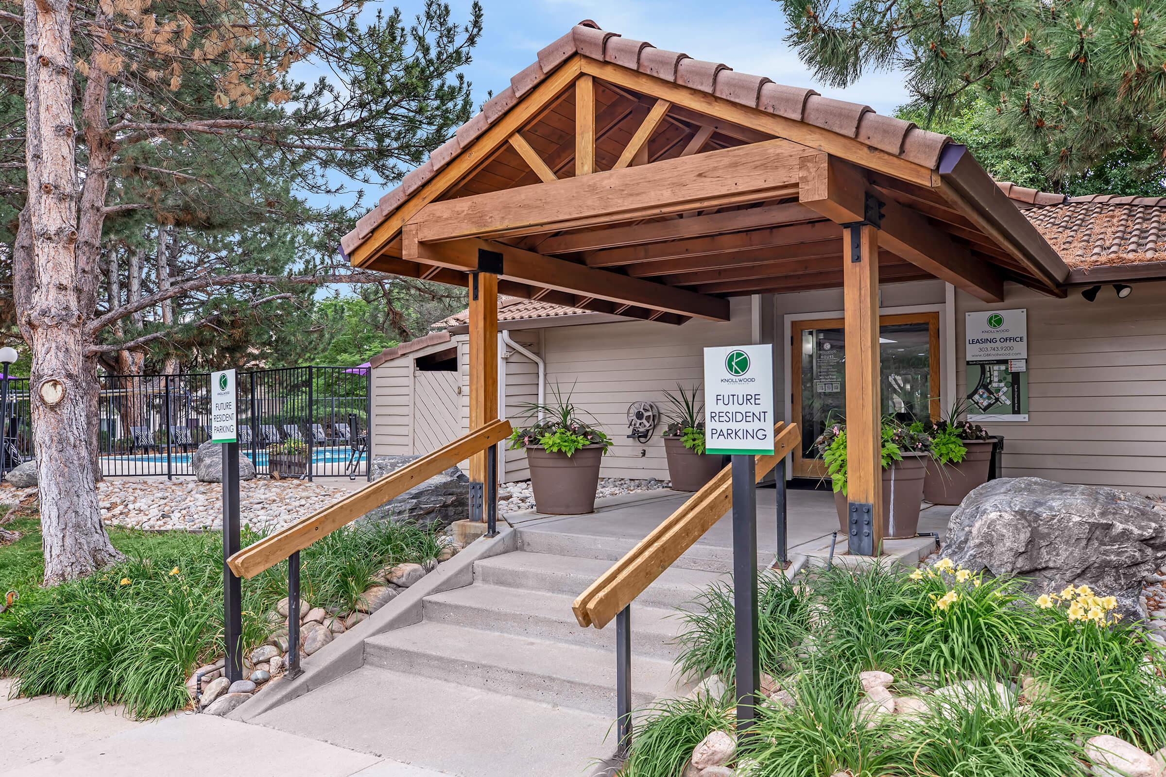 Entrance to a building with a wooden roof and steps. Signs indicate "Future Resident Parking" and "Leasing Office." The area is landscaped with greenery, plants in pots, and features a pool visible in the background behind a fence. Trees surround the entrance, creating a welcoming atmosphere.