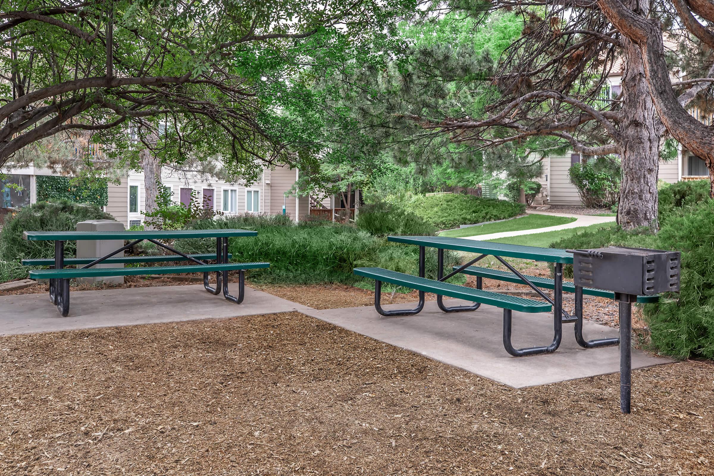 Two green picnic tables with benches are placed on a concrete pad surrounded by grass and trees. A small black grill is positioned next to one of the tables. The area appears to be a public park or recreational space, with residential buildings visible in the background.