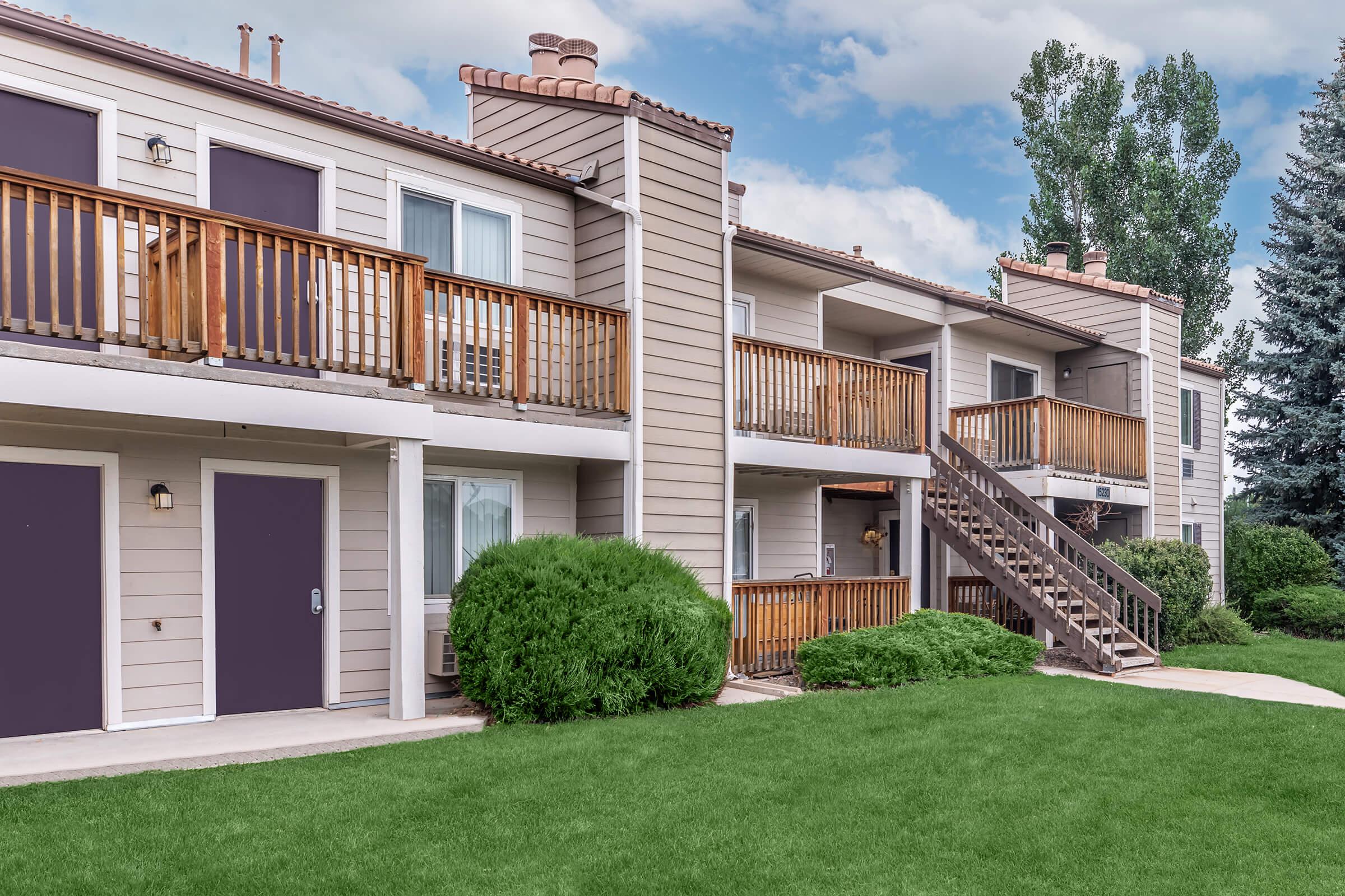 Exterior view of a two-story apartment complex featuring balconies with wooden railings, neatly trimmed bushes, and well-maintained grass. The building has light-colored siding, purple doors, and a staircase leading to the upper level. A blue sky with scattered clouds is visible in the background.