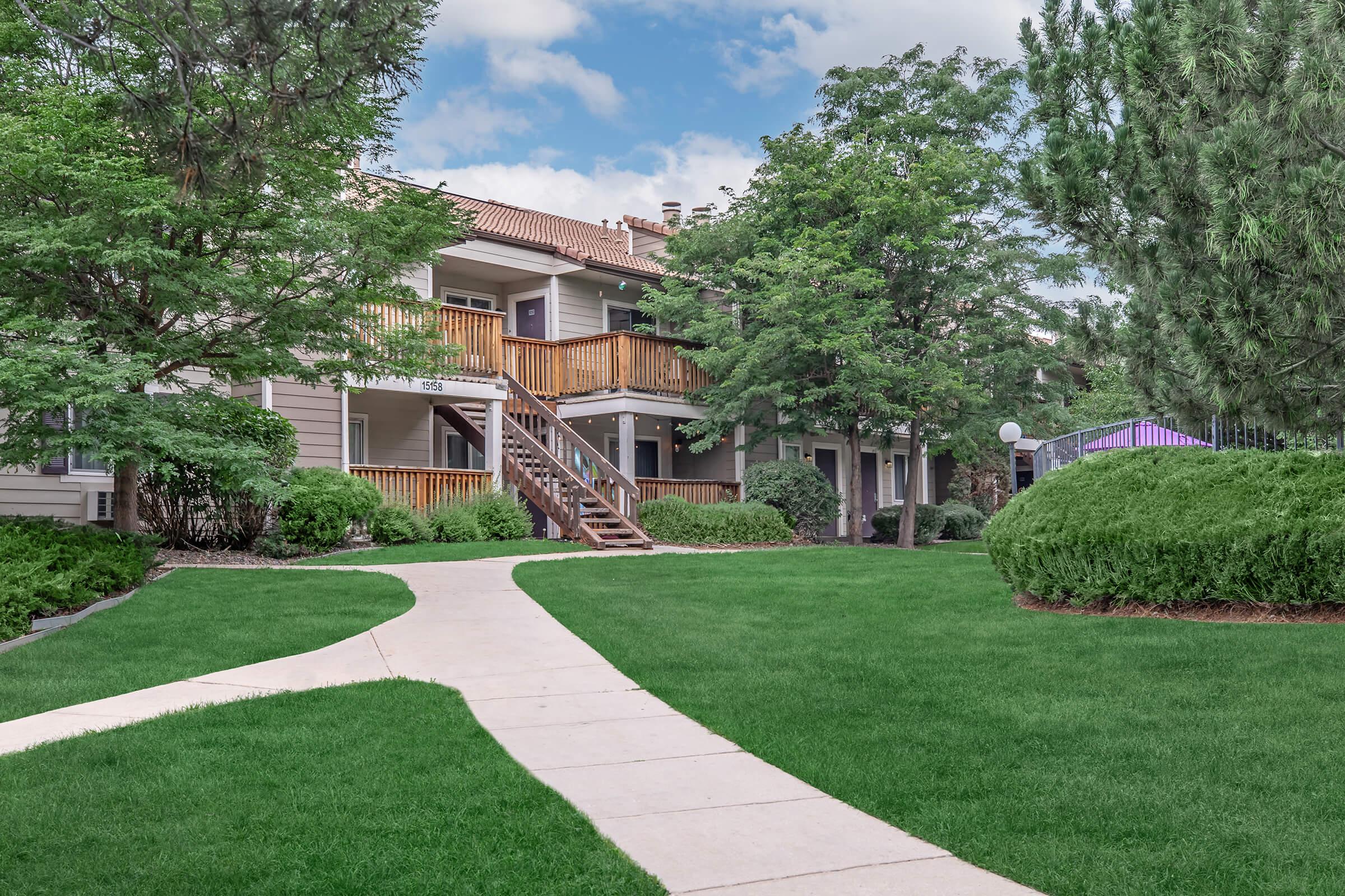 A well-maintained apartment complex surrounded by lush green lawns and trees. The path leads to two-story buildings with wooden balconies. Bright blue sky with scattered clouds adds to the inviting atmosphere.
