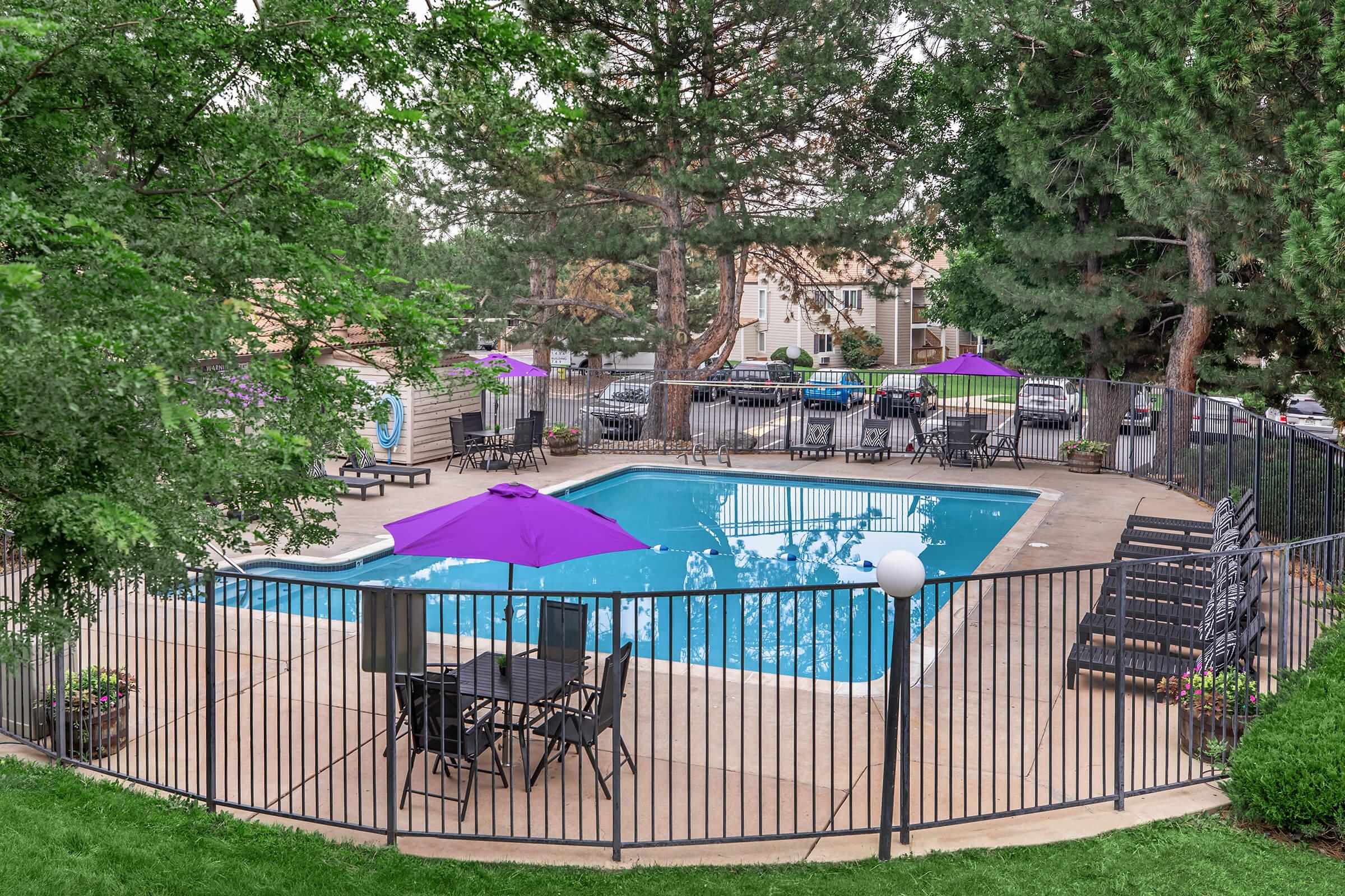 A fenced pool area featuring a blue swimming pool surrounded by green trees. There are several lounge chairs and tables with purple umbrellas for shade. In the background, parked cars are visible, and the scene is bright and inviting, suggesting a recreational space.