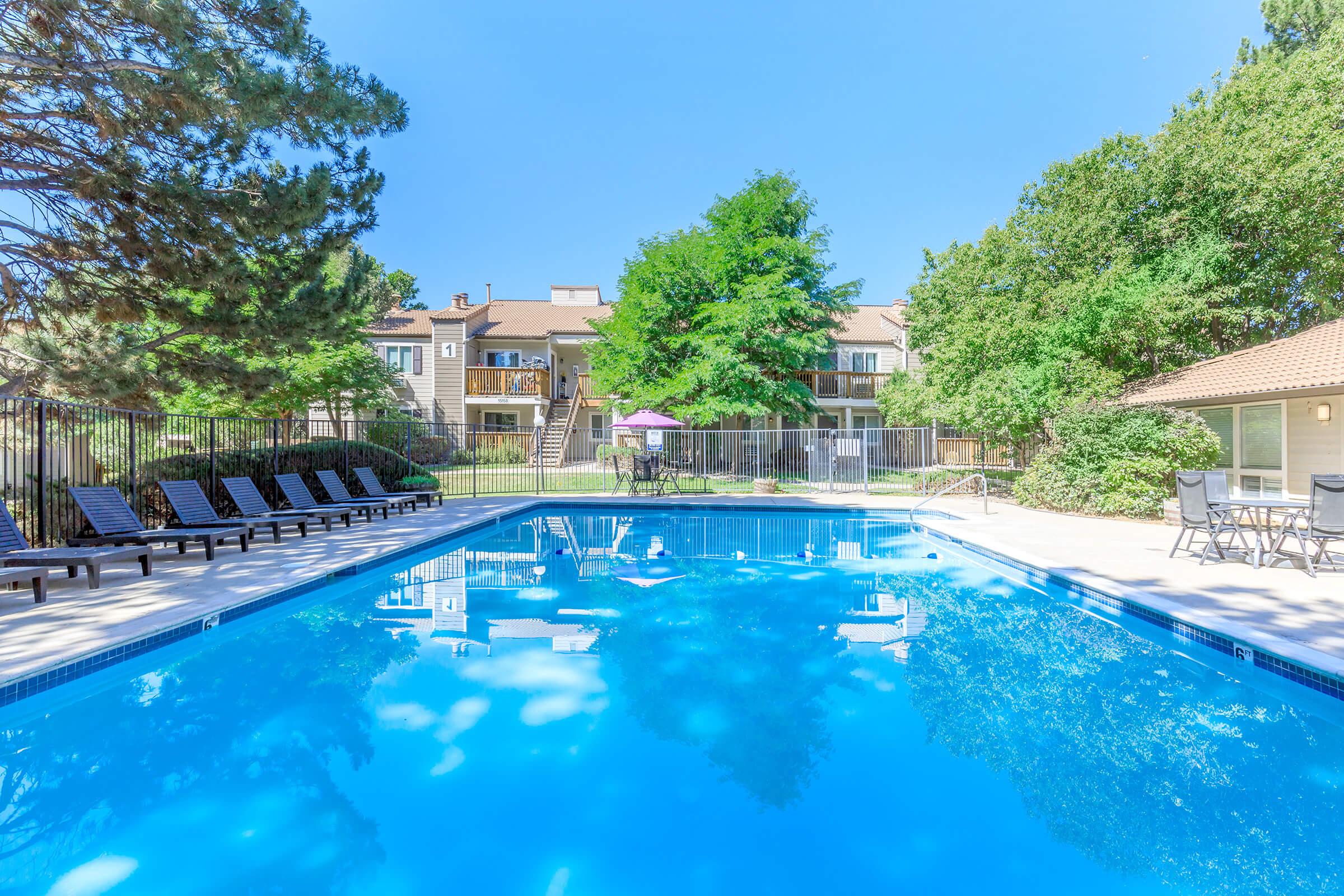 A clear blue swimming pool surrounded by lounge chairs, with lush green trees and a well-maintained lawn in the background. Apartment buildings are visible in the distance, and a purple umbrella offers shade near the pool area, creating a relaxing outdoor atmosphere on a sunny day.