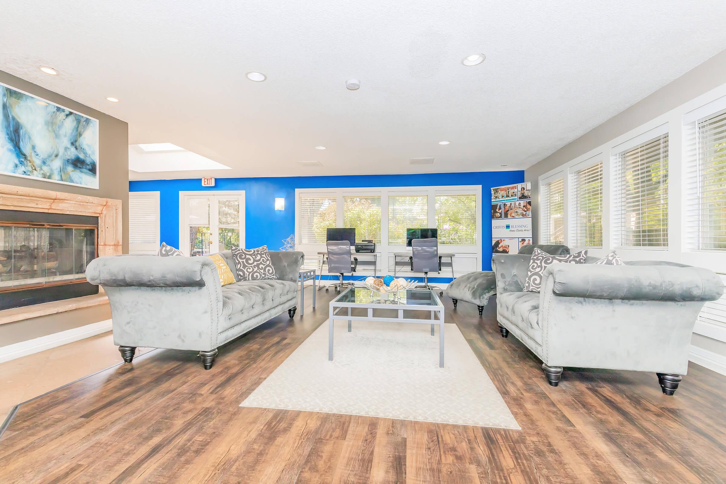 A spacious modern living area featuring two plush gray sofas, a coffee table, and bright blue accent wall. Large windows provide natural light, and there’s a cozy fireplace on one side. The decor includes decorative pillows and a rug, creating a welcoming atmosphere for relaxation and gatherings.
