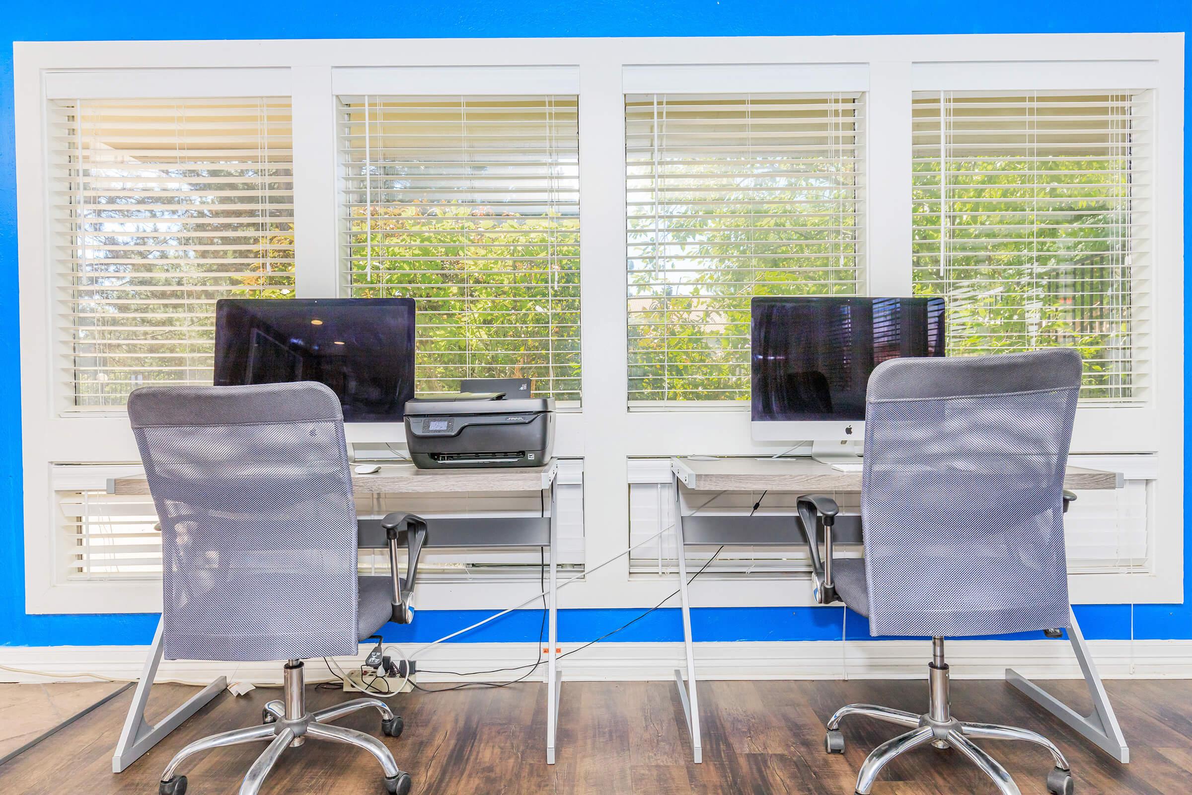 Two modern workstations facing a window with blinds, featuring two desktop computers, a printer, and ergonomic chairs. The walls are painted blue, and there is natural light coming through the window, illuminating the workspace. The floor is a dark wood finish.