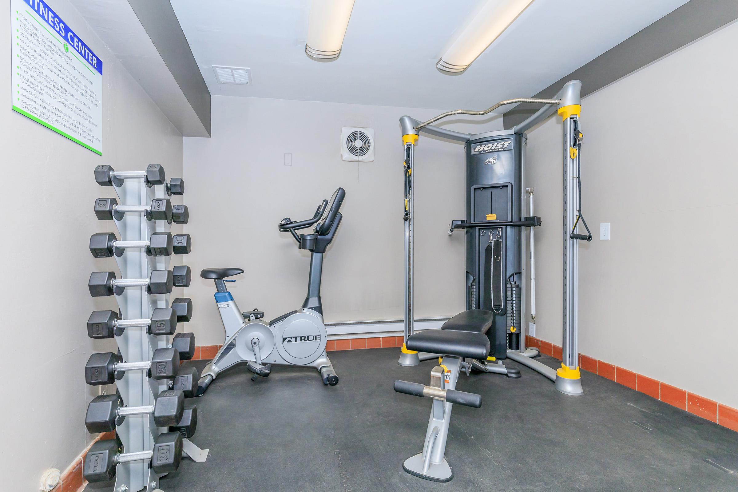 A small home gym with a multi-station workout machine, exercise bike, and a rack of dumbbells. The room features grey walls, a black floor, and overhead lighting, creating a bright and functional workout space. A fitness center information sheet is visible on the wall.