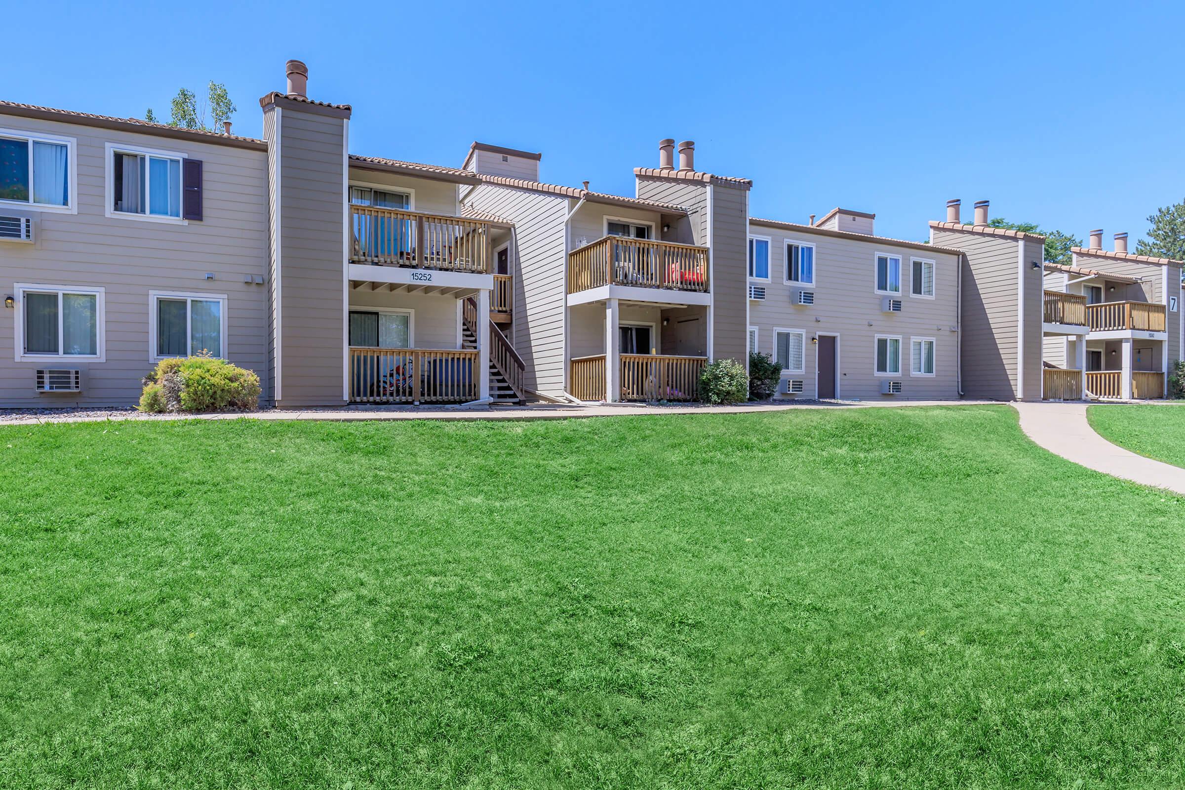A row of tan apartment buildings with balconies, set against a bright blue sky. The well-maintained lawn in front features green grass and a curved pathway leading to the doors of the apartments. Air conditioning units are visible under some windows.