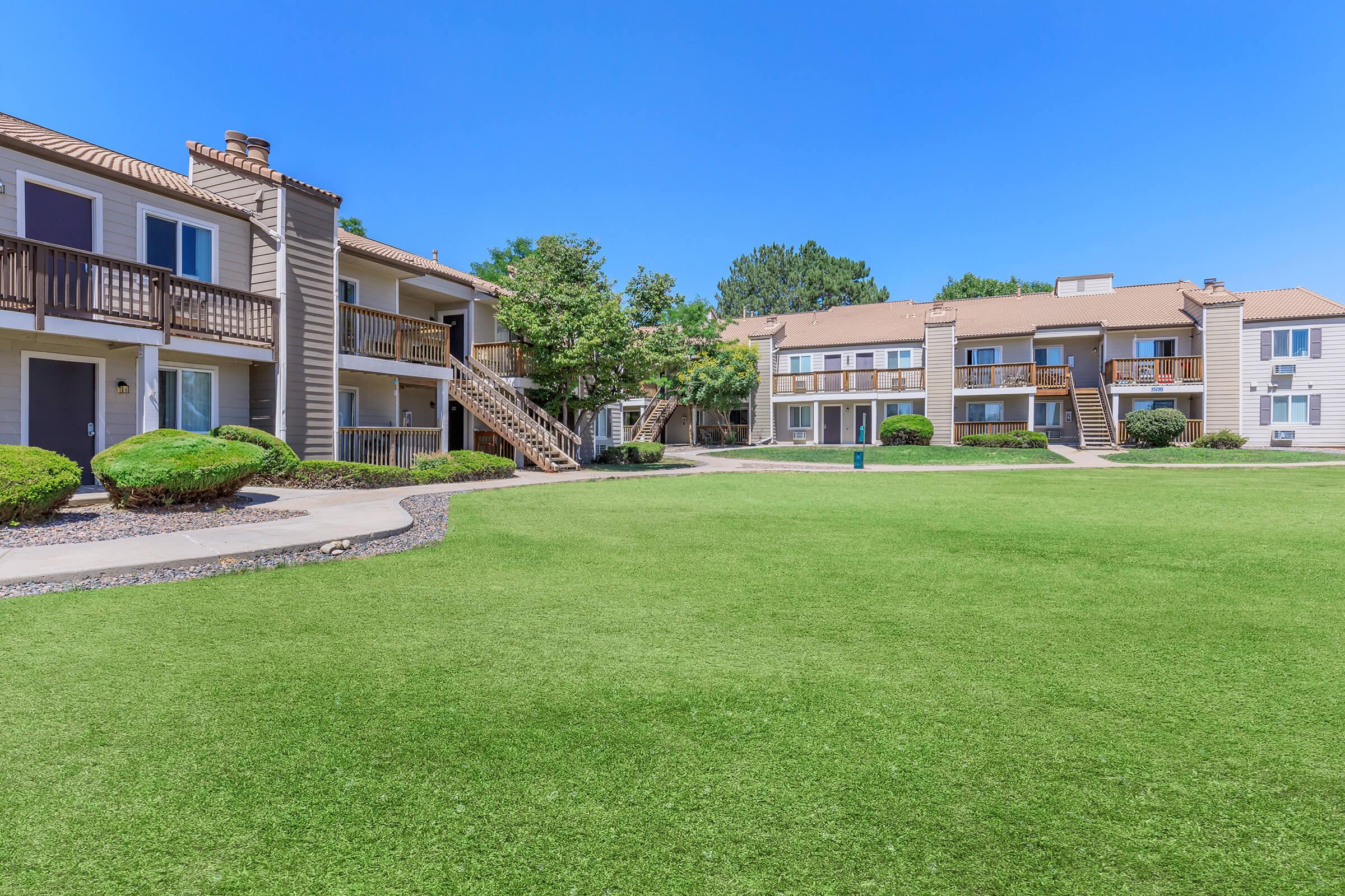 A well-maintained apartment complex featuring two buildings with balconies, surrounded by green lawns and manicured bushes under a clear blue sky. The pathway leads to the front entrances of the apartments, providing a welcoming outdoor space.