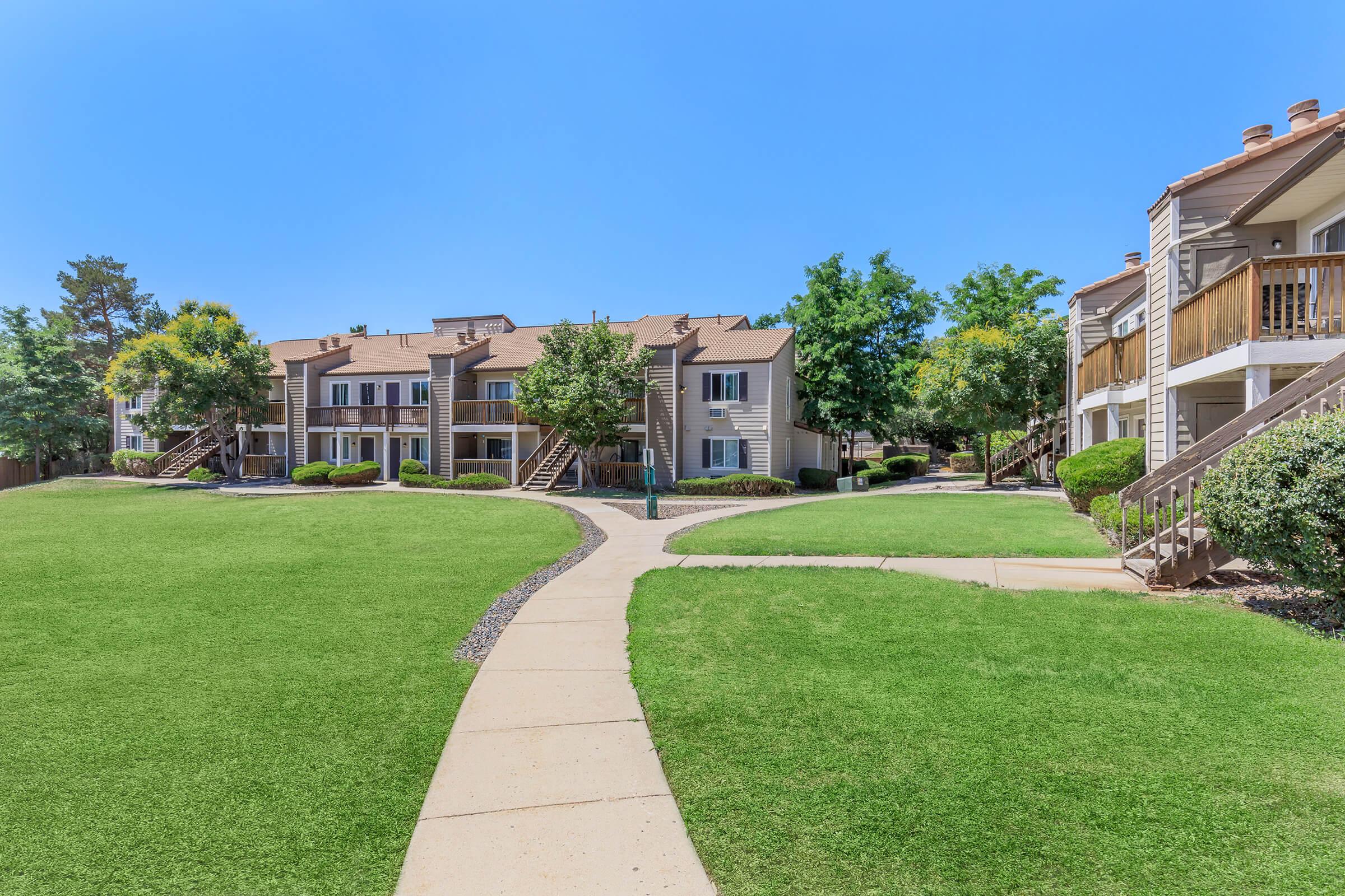 A well-maintained apartment complex with multiple buildings surrounded by lush green lawns and trees. A paved path winds through the landscape, leading to the entrances of the apartments, under a clear blue sky. The area appears inviting and well-kept, ideal for outdoor relaxation.