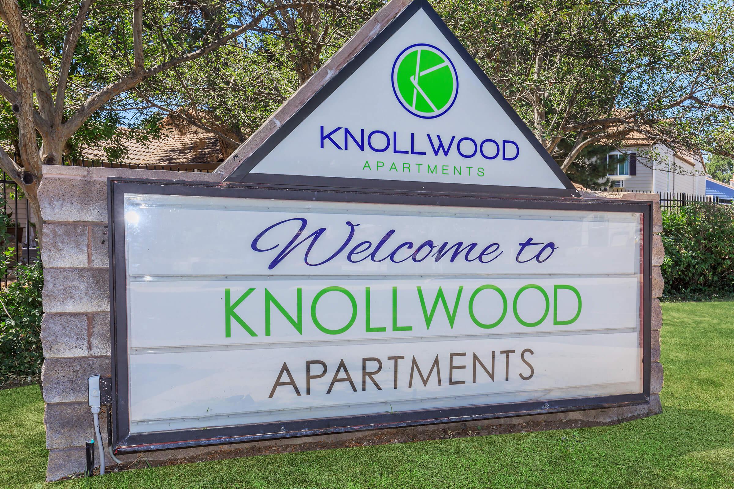 Welcome sign for Knollwood Apartments, featuring the name prominently displayed in green and blue text. The sign is set in a landscaped area with trees, and has a triangular shape at the top.