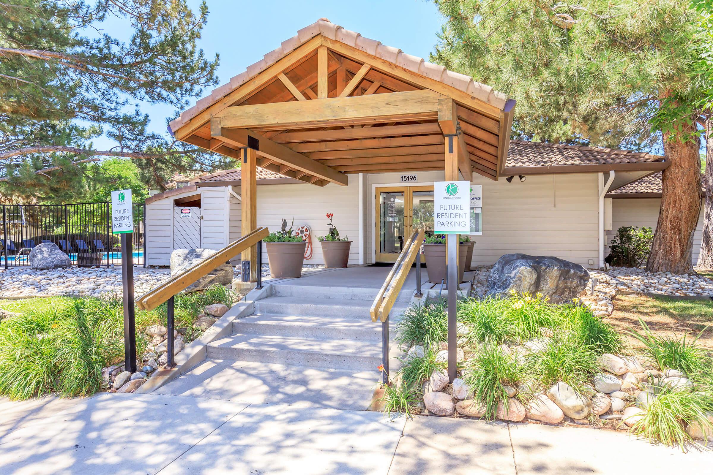 Entrance to a building with a wooden awning and steps leading up to the door. The area is landscaped with rocks and plants. Signs indicate "Future Resident Parking" on either side of the path. Trees provide shade in the background. Bright and inviting atmosphere.
