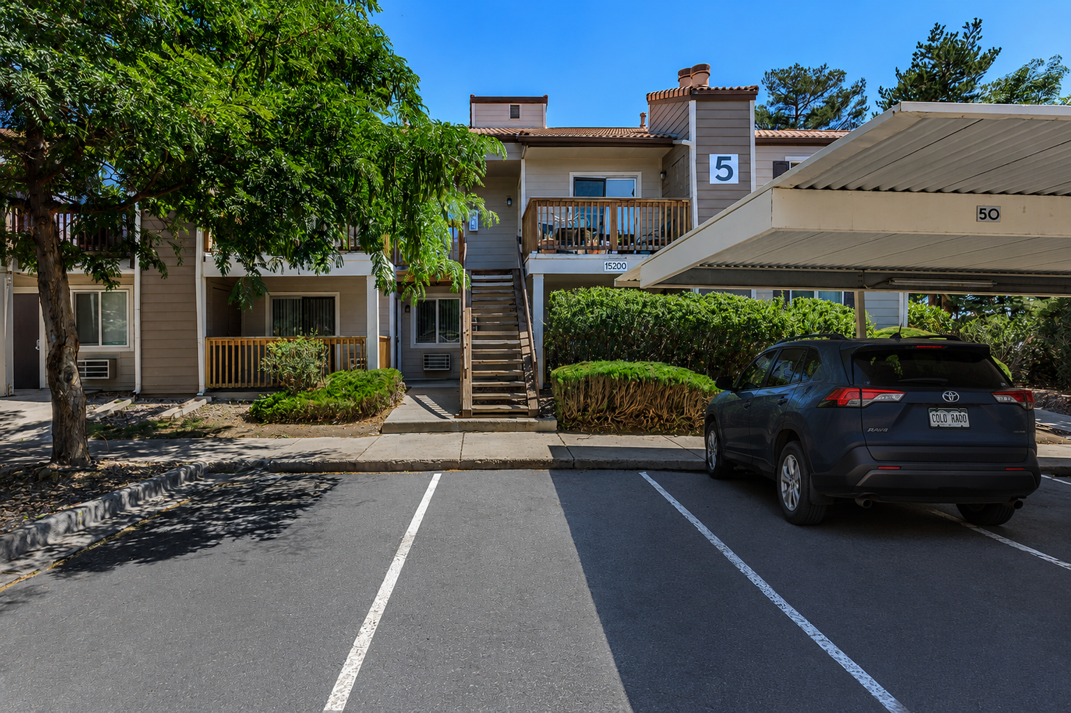 A residential apartment complex featuring a staircase leading to a second-floor unit labeled "5." In the foreground, a parking area with marked spaces, including one occupied by a dark gray SUV. Surrounding the building are trees and hedges, creating a green environment. Clear blue sky above.