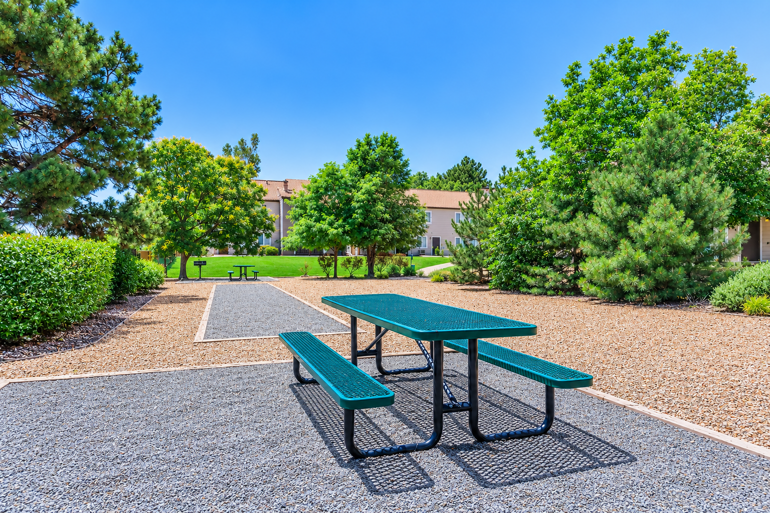 A green picnic table with attached benches in a landscaped park area featuring gravel pathways, shrubs, and trees under a clear blue sky. In the background, there are buildings partially obscured by greenery.
