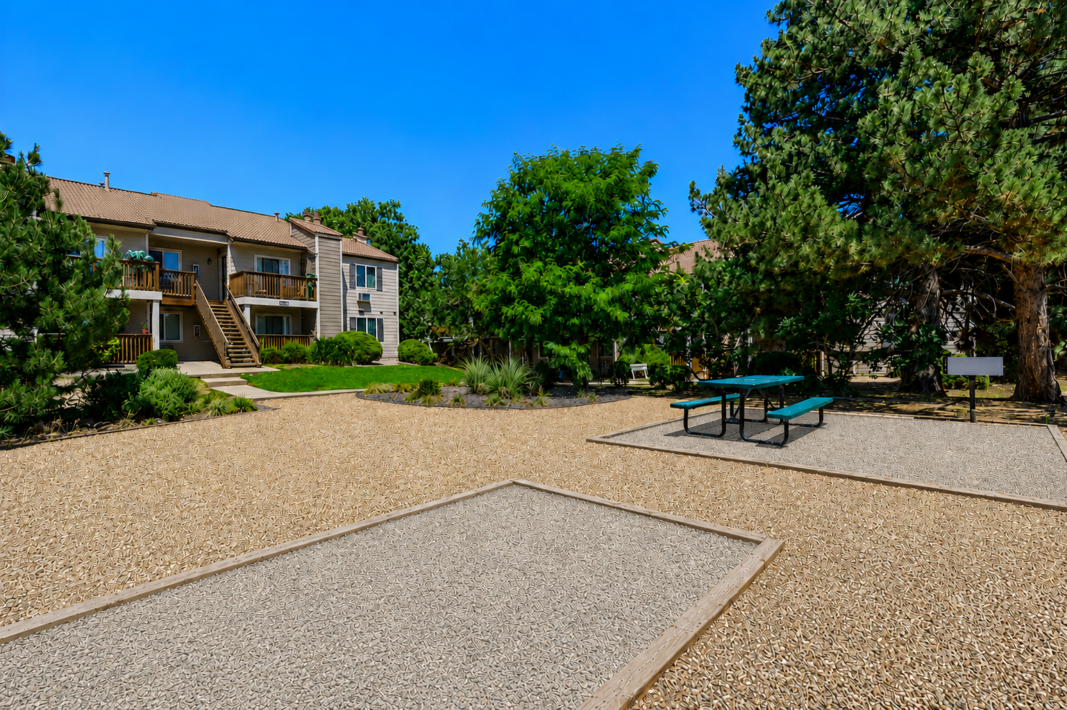 A well-maintained outdoor area featuring a gravel surface, surrounded by greenery and trees. In the background, there are two residential buildings with balconies. A picnic table is set in the foreground, providing a space for outdoor gatherings. The sky is clear and blue, emphasizing a sunny day.