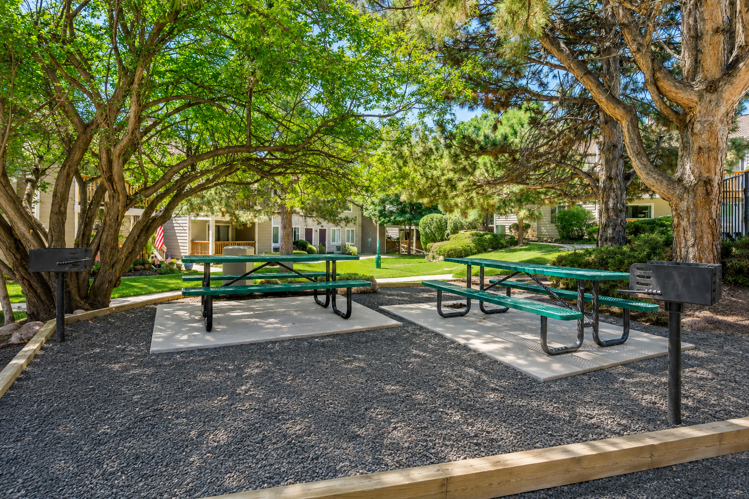 Two picnic tables with green tops are situated on a gravel area surrounded by trees in a landscaped outdoor space. The setting features well-maintained grass and shrubbery, suggesting a communal or recreational area, ideal for gatherings or relaxation.