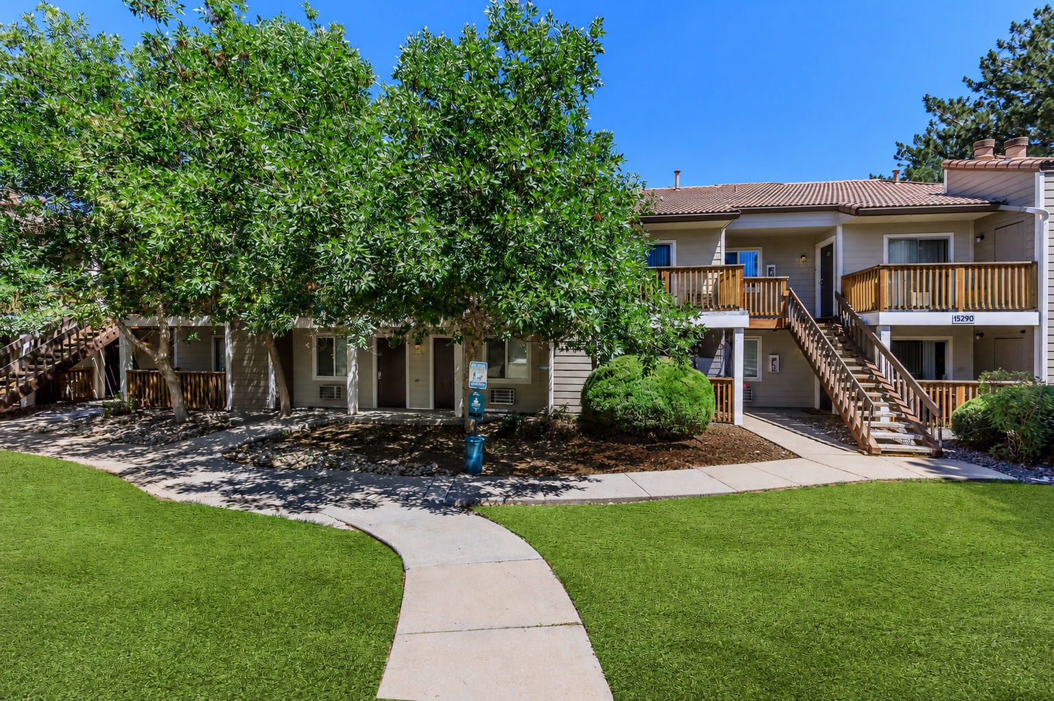 A well-maintained residential complex featuring two-story buildings with wooden balconies. Lush greenery surrounds the area, with trees providing shade. A pathway curves through a manicured lawn, leading to the entrance of the buildings. Clear blue skies create a bright and inviting atmosphere.