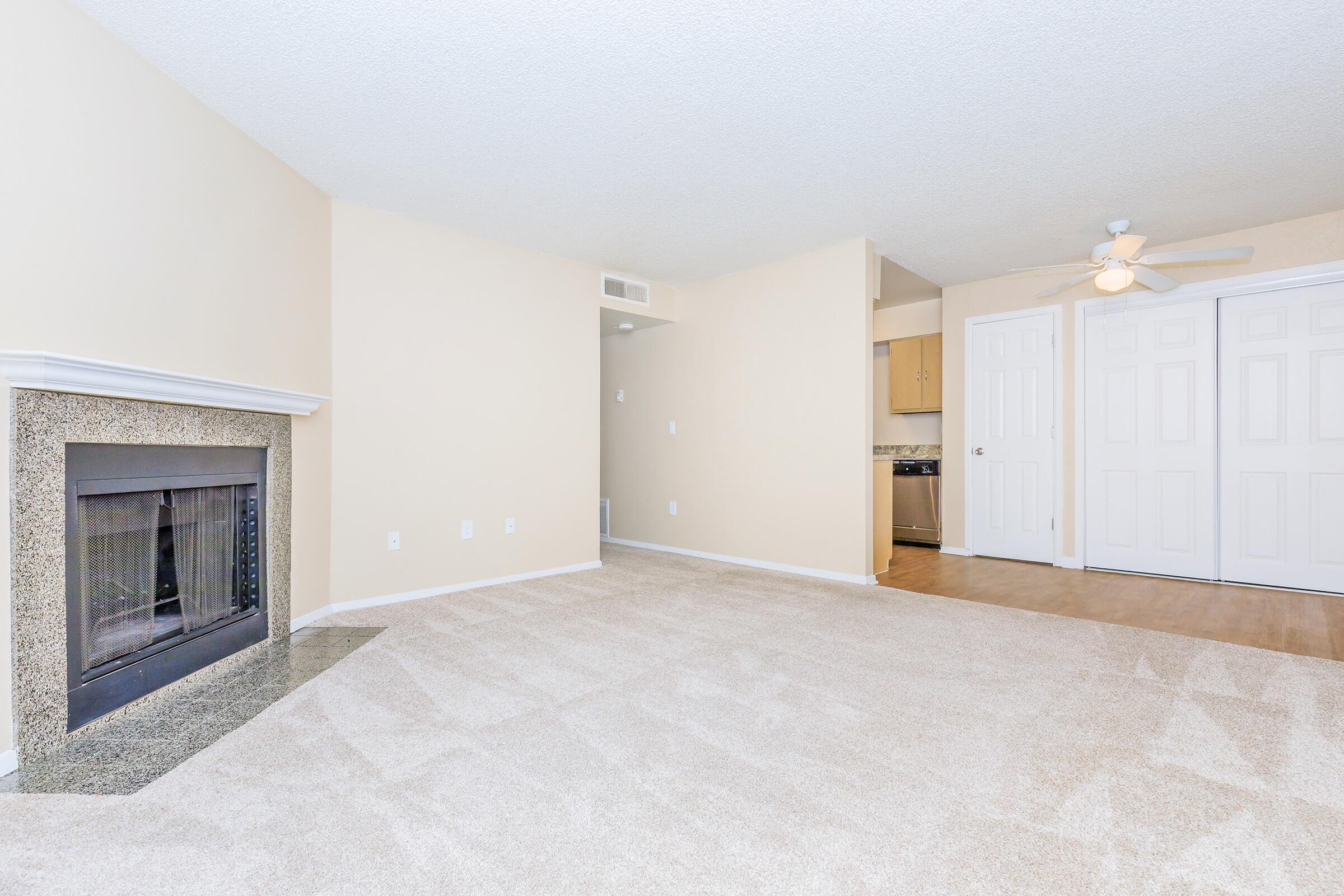 A spacious living area with beige walls and carpet, featuring a decorative fireplace on one side. To the right, there are double doors leading to a storage area or closet. In the background, a glimpse of a kitchen area is visible, with light-colored cabinetry. The room is well-lit and inviting.