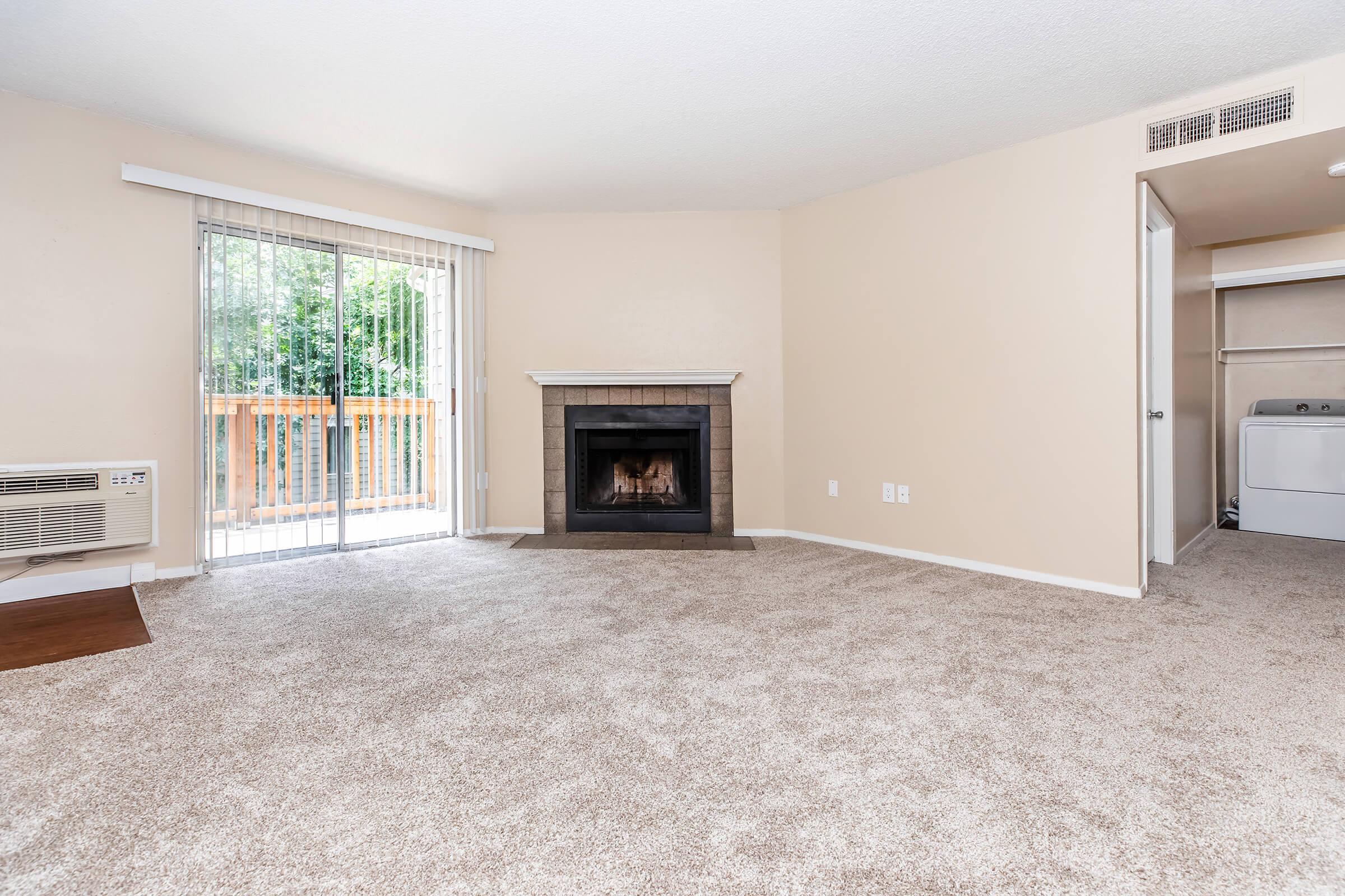 A spacious, light-filled living room with beige walls and plush carpet. It features a fireplace with a dark surround, large windows with sliding glass doors leading to a balcony, and an air conditioning unit. A laundry area is visible through an open door in the background.
