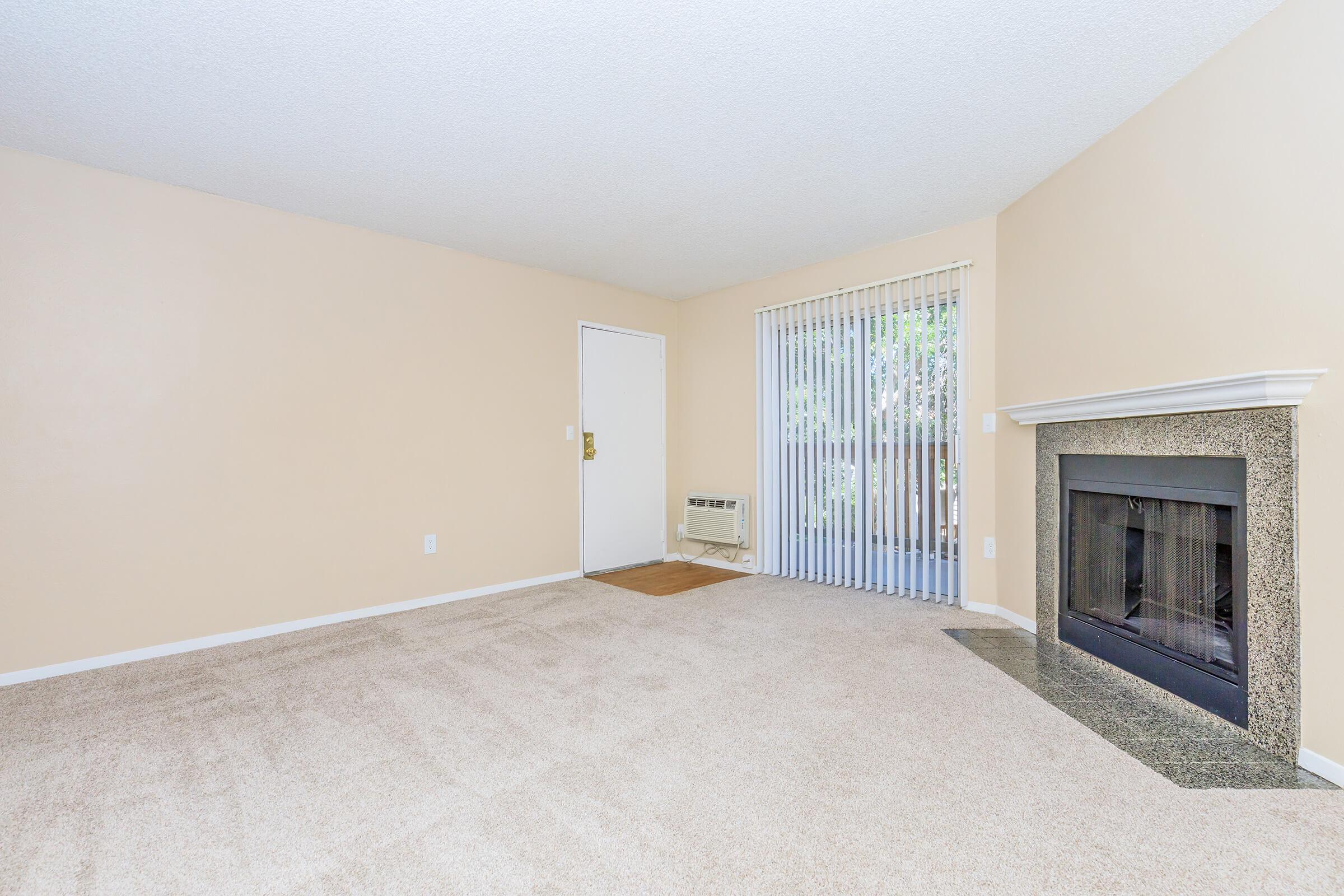 A spacious, empty living room featuring beige walls and carpet. On one side, there is a closed door, and adjacent to it, a large window with vertical blinds letting in natural light. A decorative fireplace is located along one wall, adding a cozy element to the room.