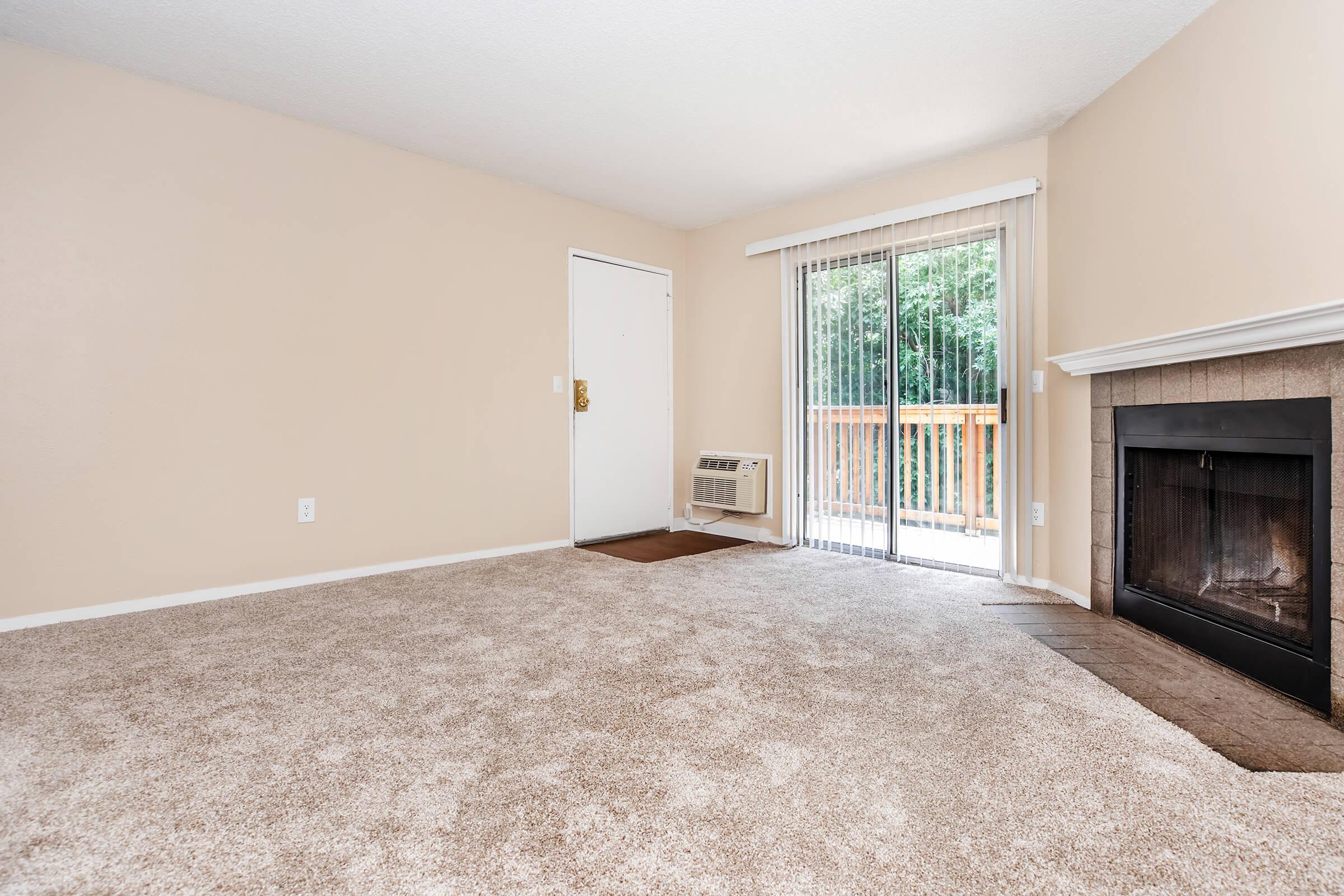 Empty living room with beige walls and carpet. A door leads outside, with a sliding glass door adjacent to it, allowing natural light. An air conditioning unit is installed below the window. The room features a simple fireplace with a tiled surround, creating a cozy atmosphere.