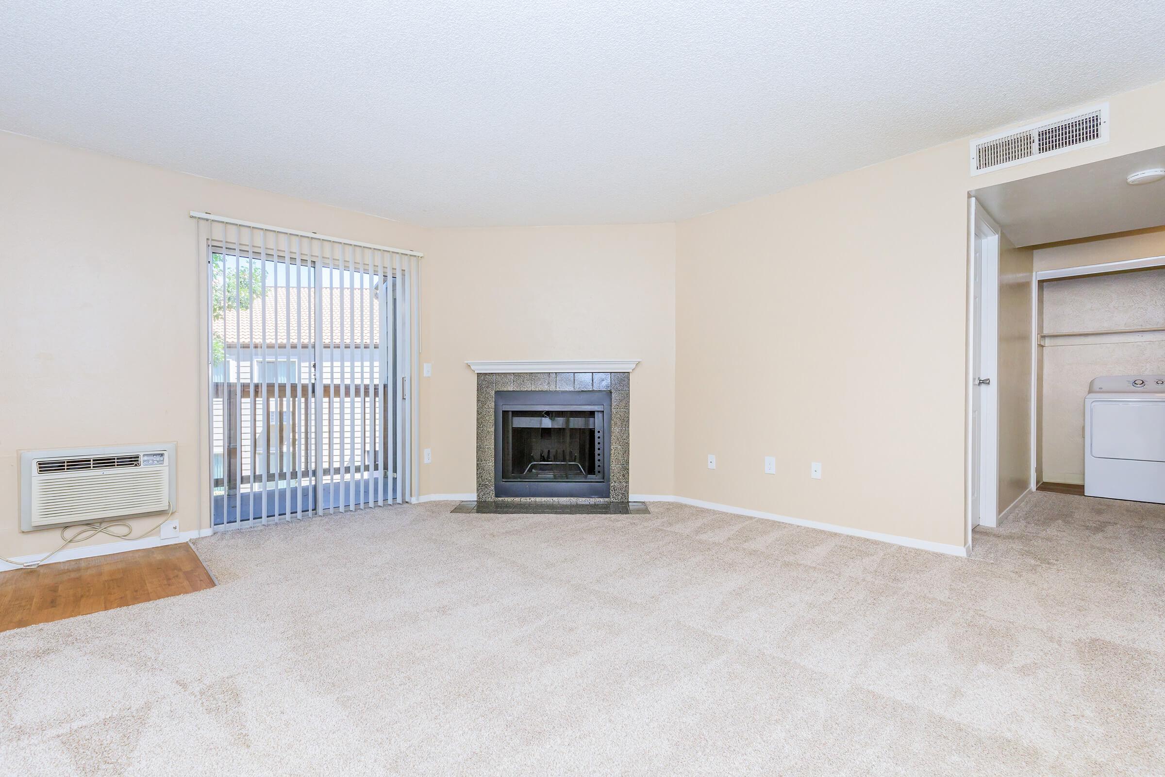 Spacious living area with beige walls and carpet, featuring a decorative fireplace. A sliding glass door leads to a balcony with vertical blinds. An air conditioning unit is mounted on the wall, and there's a laundry area visible through an open door. Natural light fills the room, creating a warm and inviting atmosphere.