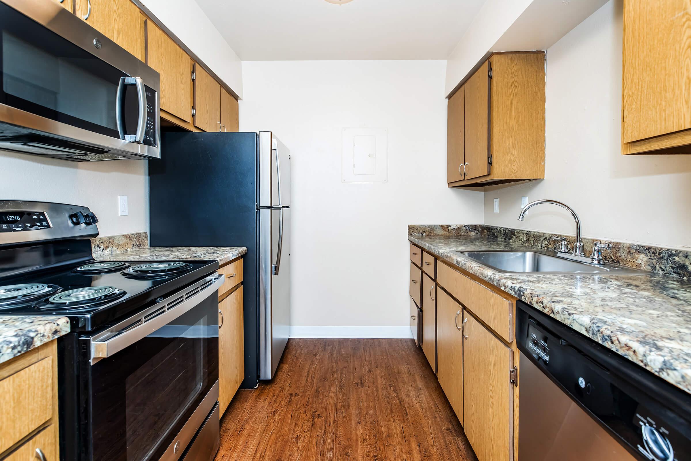 A modern kitchen featuring wooden cabinets, stainless steel appliances, a double sink, and a granite countertop. Includes an electric stove, microwave, dishwasher, and a refrigerator. The floor has a wood-like finish, creating a warm and inviting atmosphere.