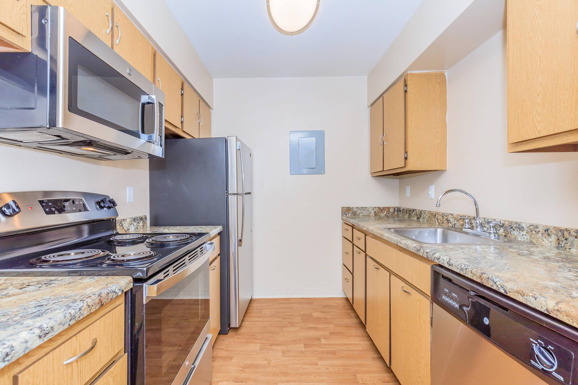 A modern kitchen featuring wooden cabinets, stainless steel appliances including a microwave, oven, and refrigerator, a sink with a countertop, and a dishwasher. The floor is made of light-colored wood, and the walls are painted in a neutral tone, creating a bright and inviting space.