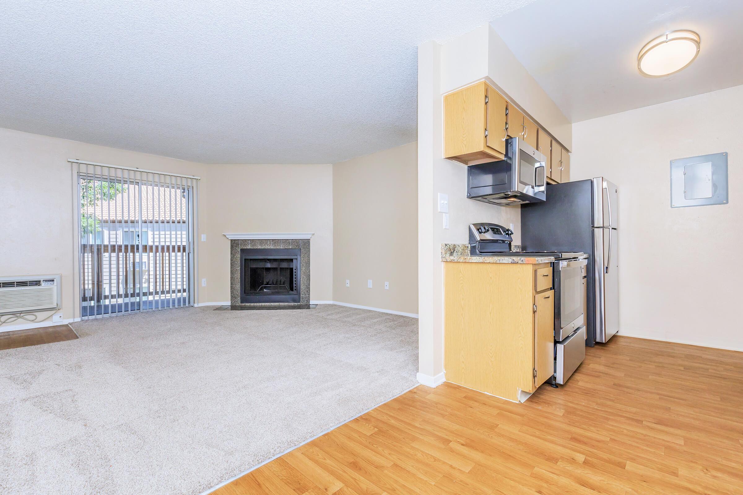 A modern apartment interior featuring a spacious living area with beige carpeting, a fireplace, and a large window with blinds. To the right is a kitchen area with wooden cabinets, a stainless steel microwave, a refrigerator, and a stove. Soft natural lighting enhances the welcoming atmosphere.