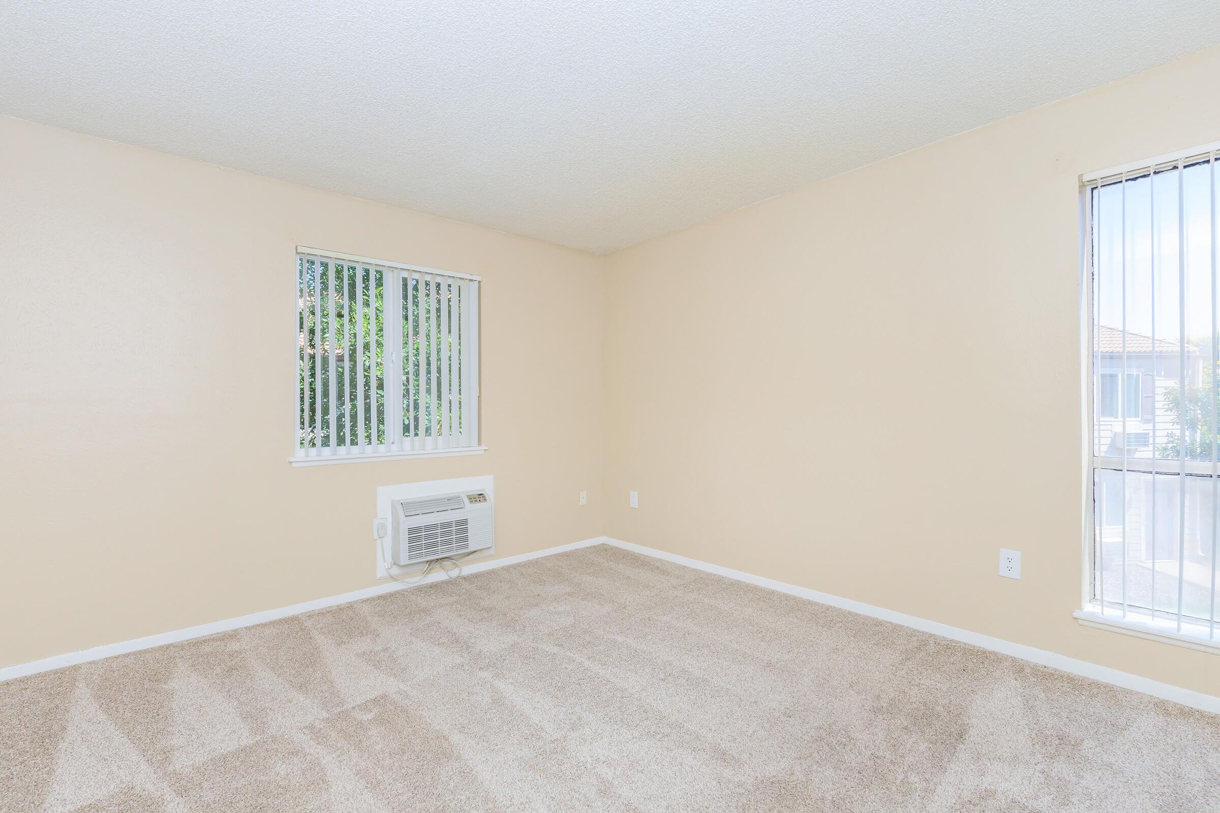 A bright, empty room with beige walls and light brown carpet. It features two windows with vertical blinds, one of which is partially open. An air conditioning unit is installed on one wall, and the overall atmosphere is spacious and inviting, perfect for personalization.
