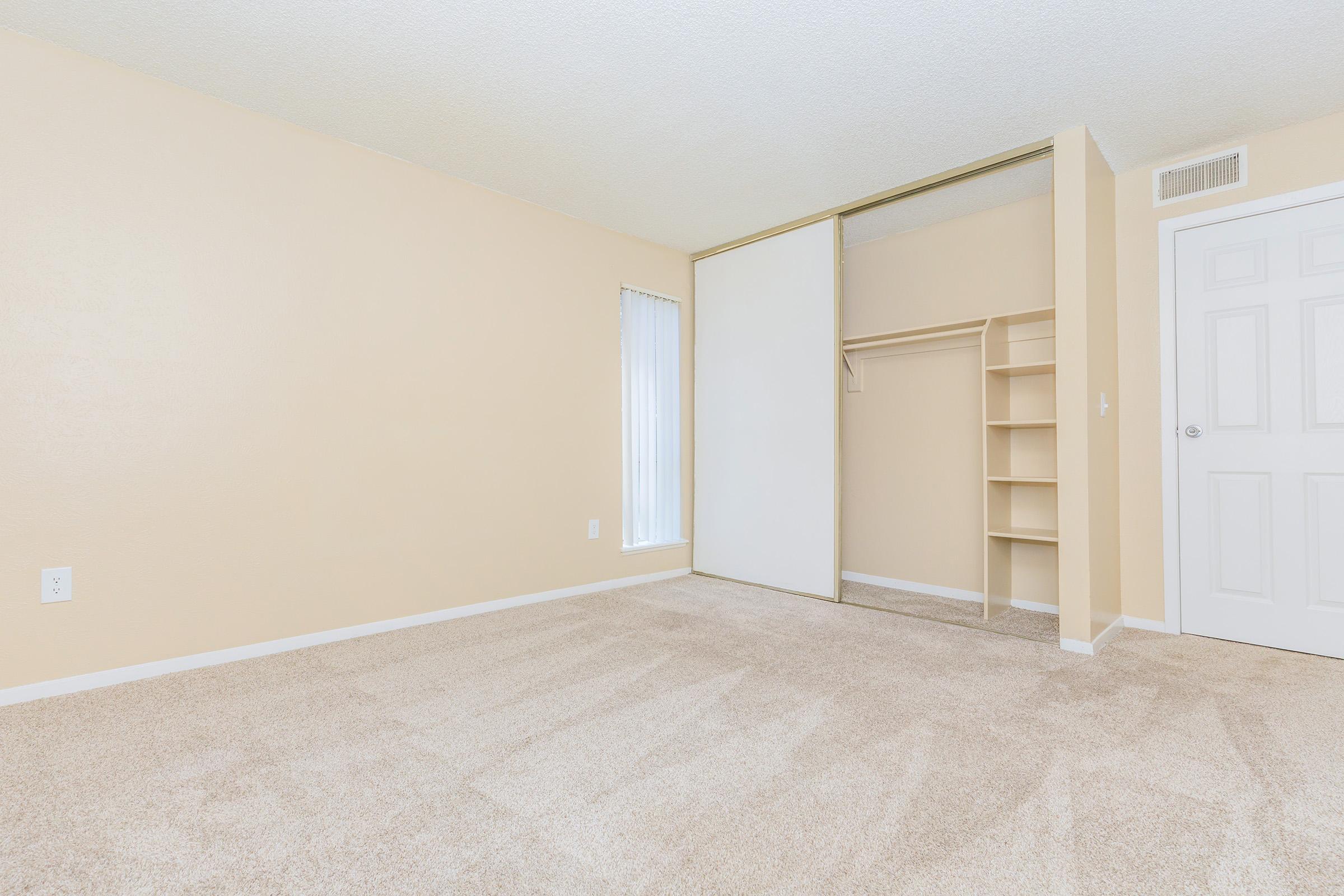 A vacant room with light beige walls and carpet. The room features a large sliding door closet with built-in shelves, a window with vertical blinds, and a white door. The space is bright and airy, showcasing a minimalist design.