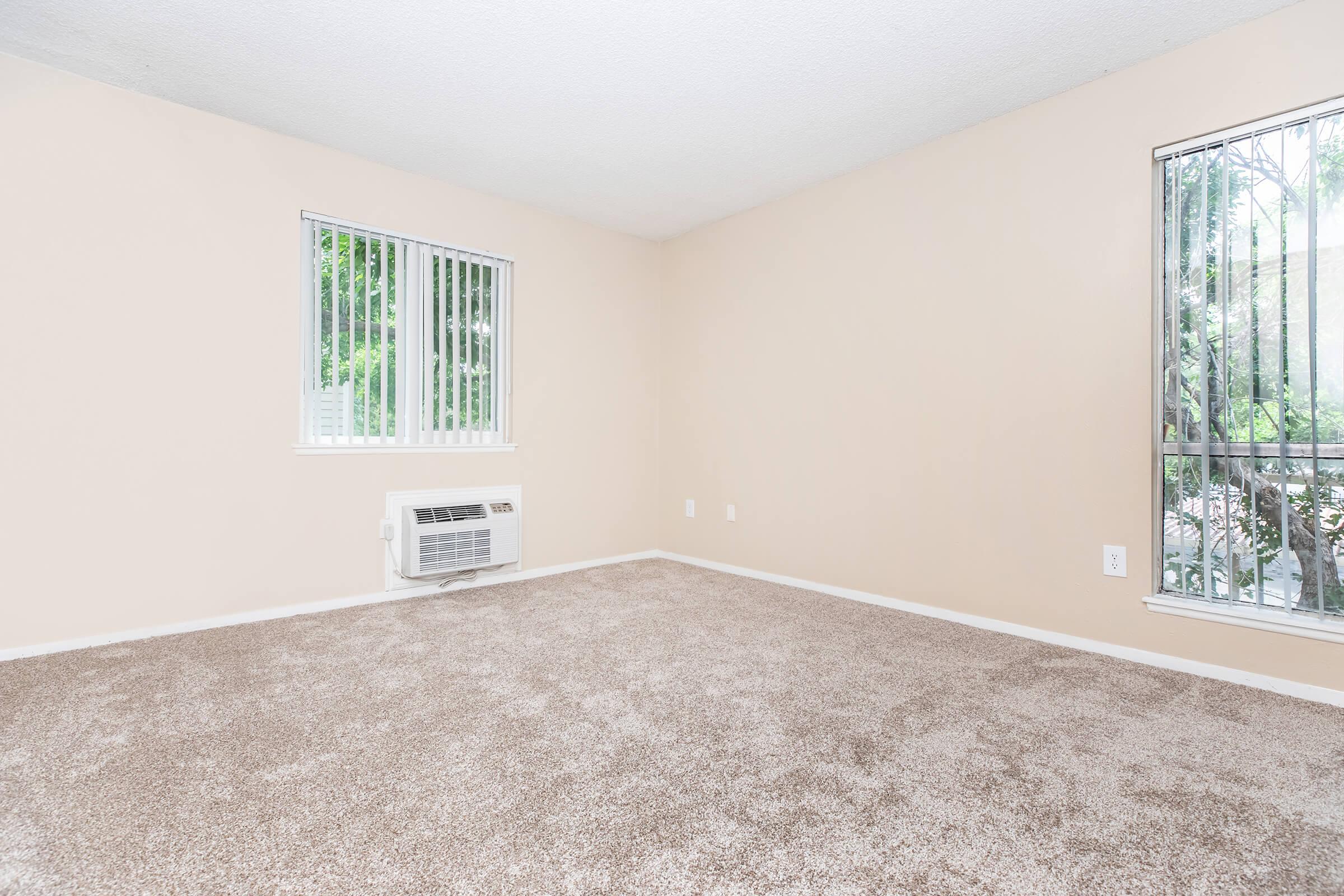 A spacious, empty room with light beige walls and carpeting. It features two windows with vertical blinds allowing natural light. An air conditioning unit is installed on one wall. The overall atmosphere is clean and minimalistic, suitable for various interior designs.