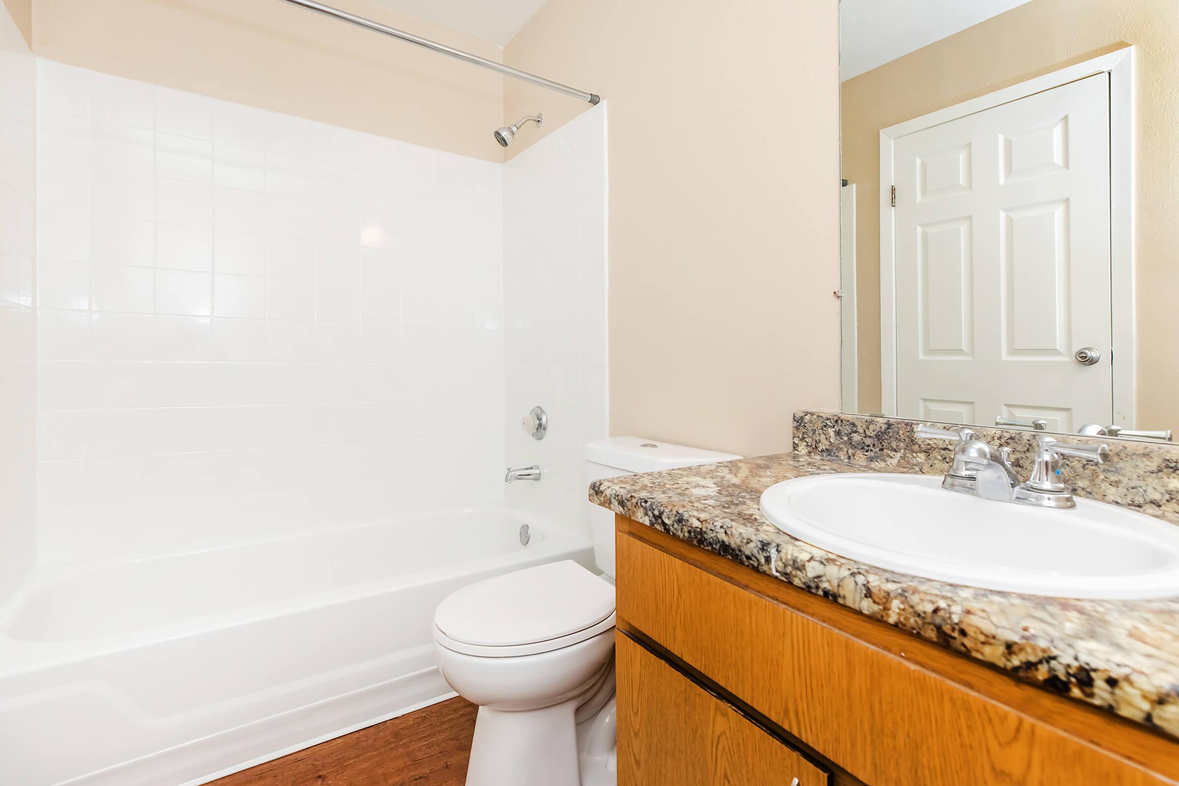 A clean and modern bathroom featuring a white tub and shower, a granite countertop sink area, wooden cabinetry, and a mirror. The walls are painted a soft beige, and there is a door leading to another room. The flooring is a light wood finish.