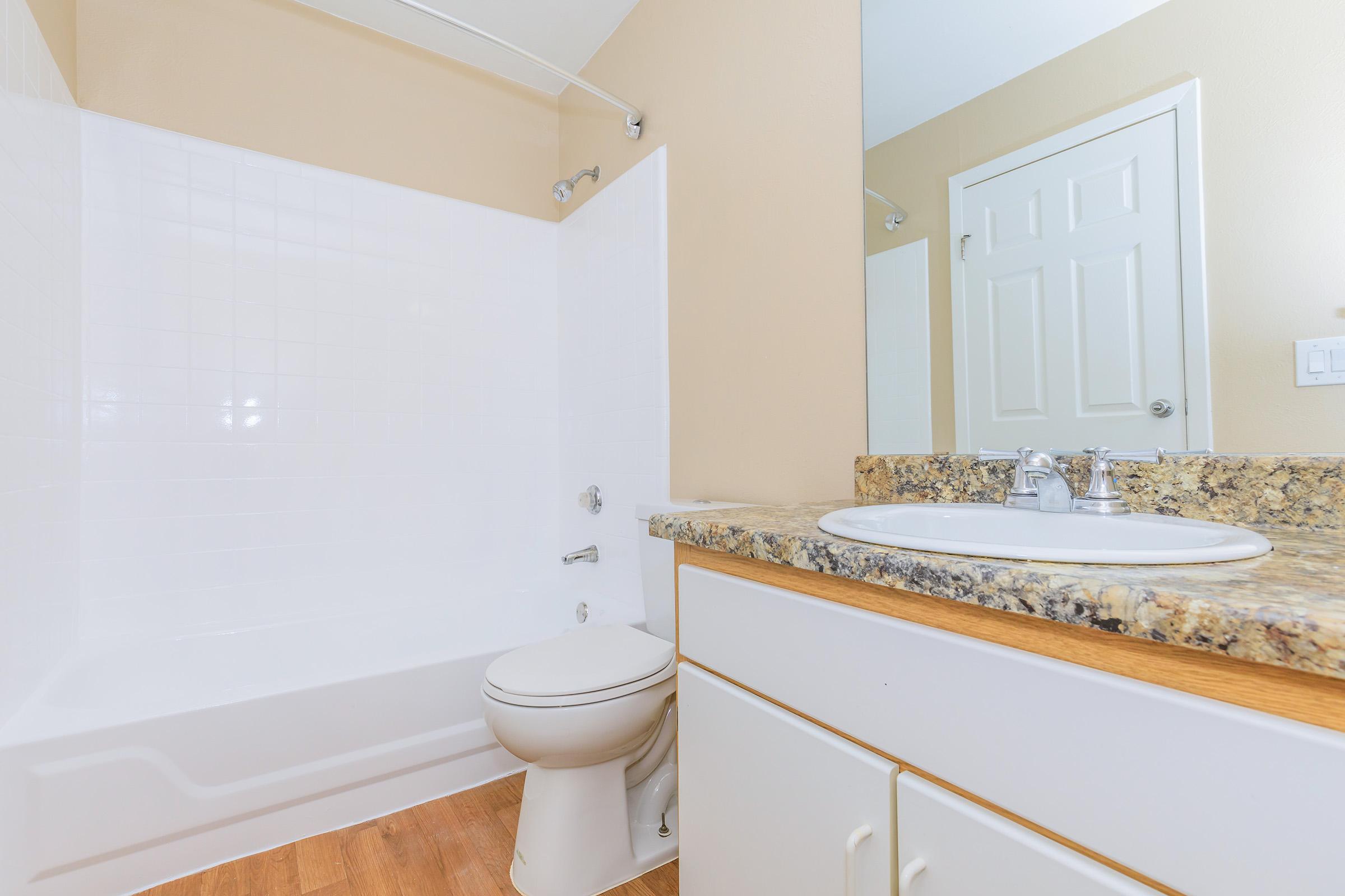 A clean, modern bathroom featuring a white bathtub and shower combination, a toilet, and a wooden vanity with a sink. The walls are painted a soft beige, and a mirror is mounted above the sink. Natural light illuminates the space, complemented by simple decor.