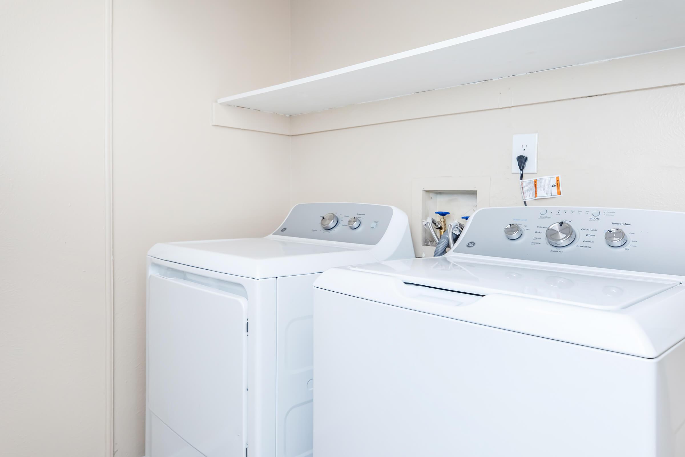 A clean laundry room featuring a white washing machine and a dryer side by side. Above them is a shelf with some items. The walls are a light neutral color, creating a simple and functional space for doing laundry.