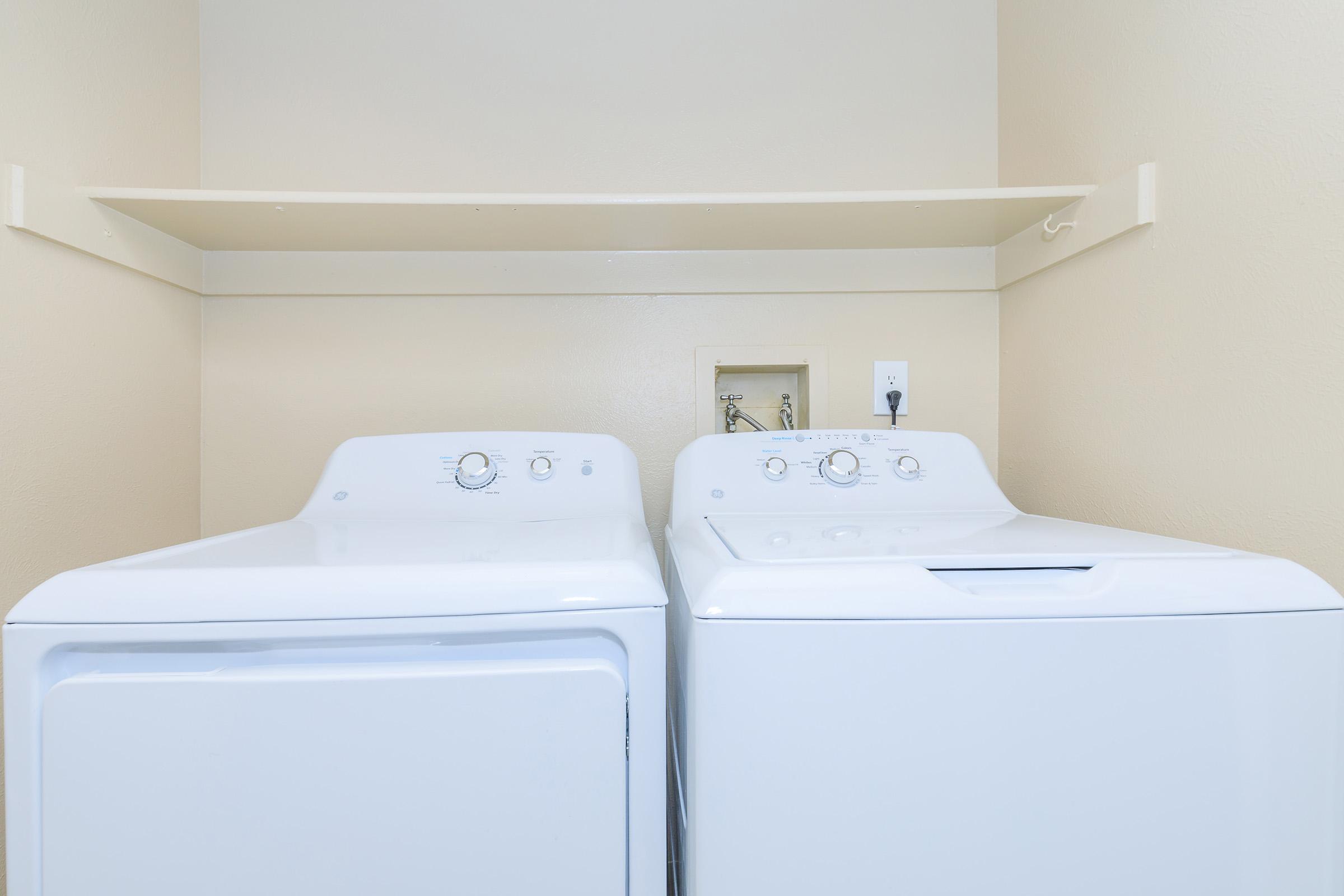Two white laundry appliances, a washing machine and a dryer, side by side in a small laundry area. The wall behind them is a light beige color, and there is a shelf above the appliances. A power outlet is visible on the wall. The scene is tidy and well-lit.