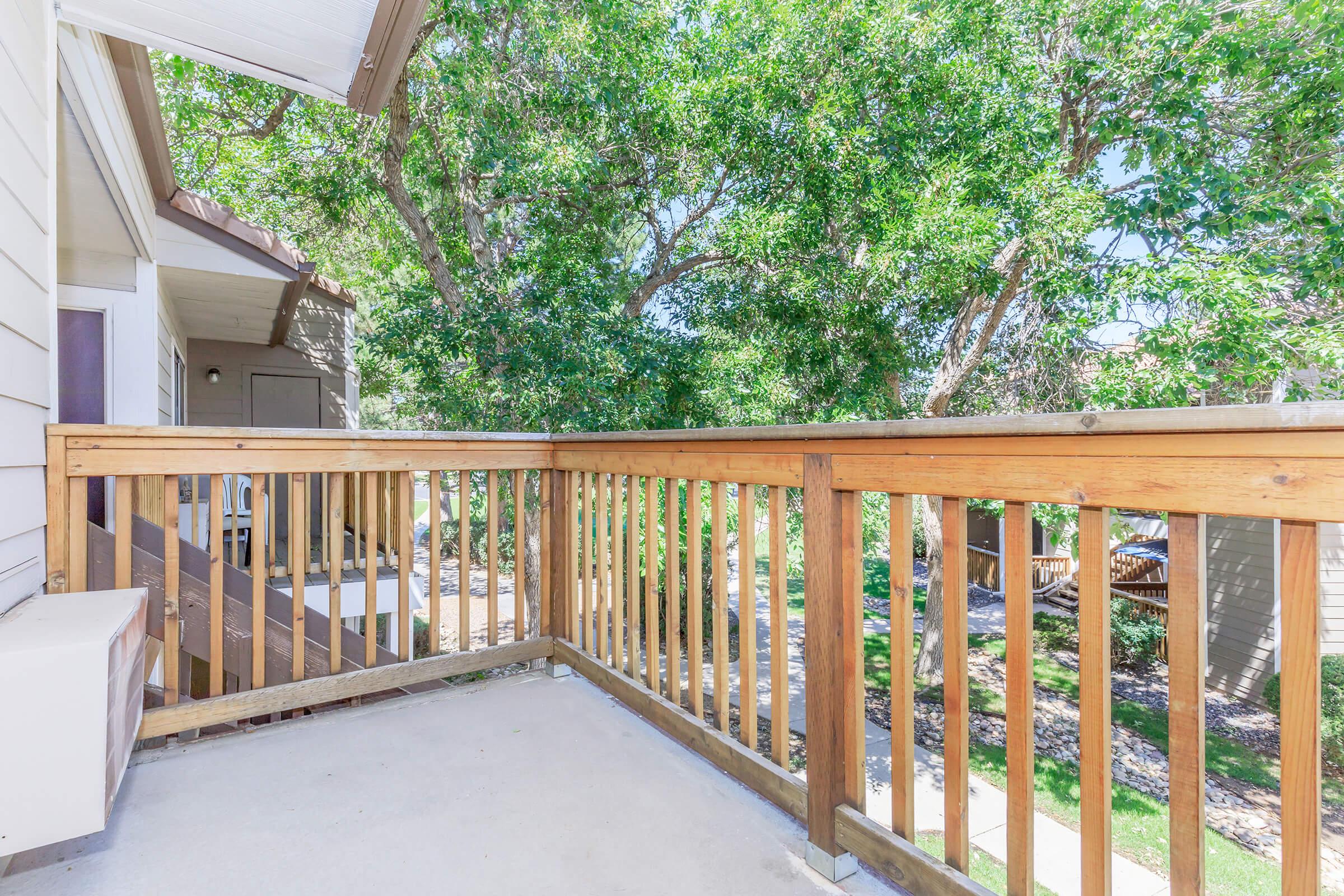 A wooden balcony railing surrounded by lush green trees, with a clear blue sky above. The balcony is part of a residential building, featuring a concrete floor and a view of the landscaped area below. Sunlight filters through the foliage, creating a bright and inviting atmosphere.