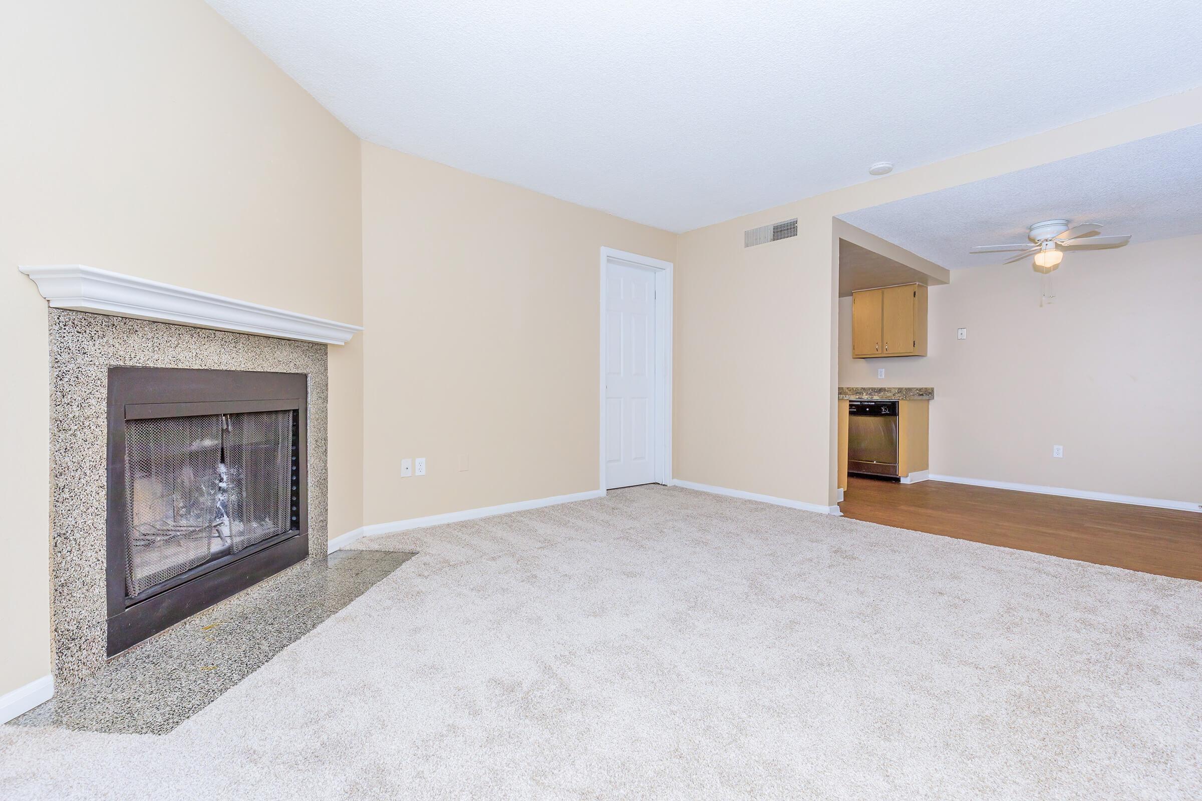 A spacious living room with beige walls and plush carpet, featuring a decorative fireplace on one wall. To the right, a doorway leads to a kitchen area with light wood cabinetry and appliances visible. The room is well-lit with natural light, creating a welcoming atmosphere.