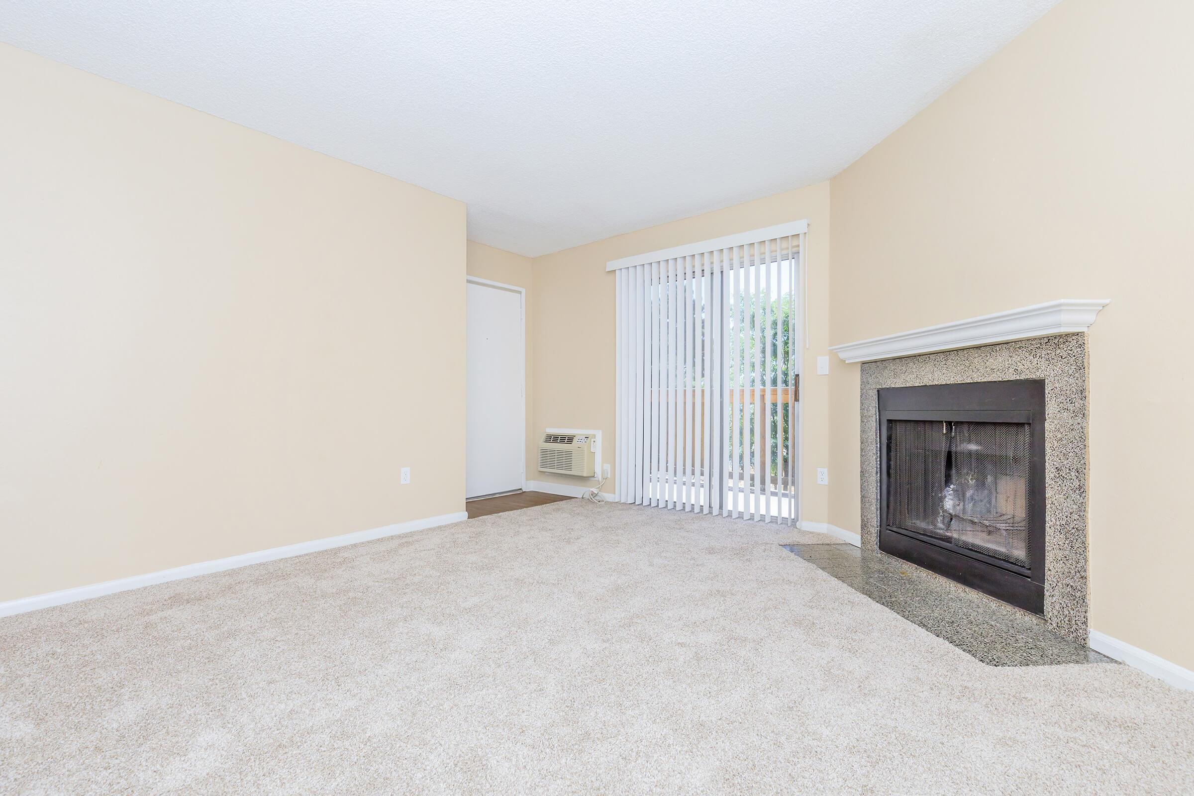 A spacious living room featuring beige walls and light carpet. There is a fireplace with a granite surround on one side, and large sliding glass doors leading to a patio. An air conditioning unit is visible near the entrance, creating a cozy and airy atmosphere.