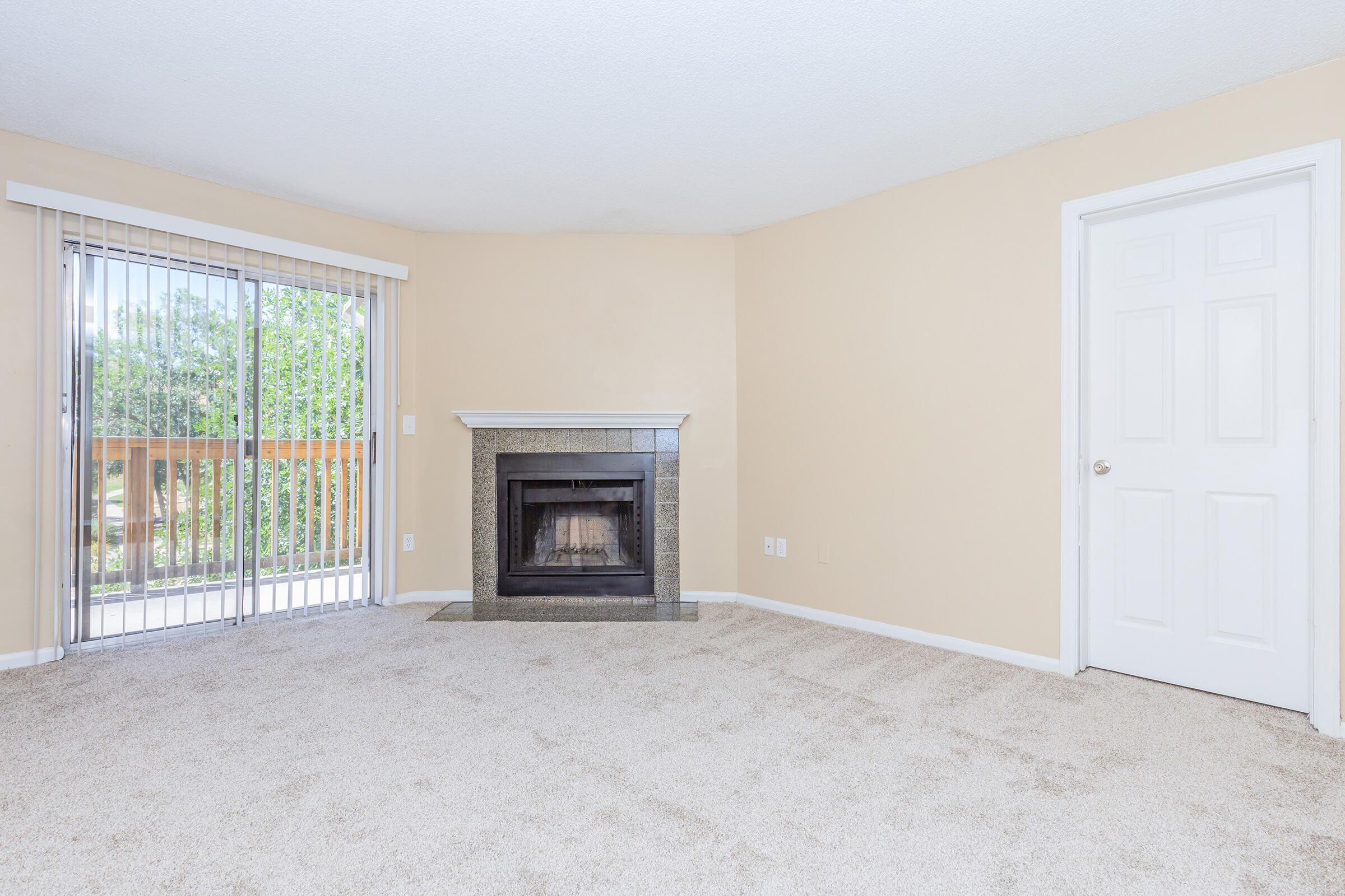 Empty living room featuring beige walls and carpet, a fireplace with a granite surround, and sliding glass doors leading to a balcony. A white door is visible on the right side. The room is well-lit, with natural light filtering through the doors.