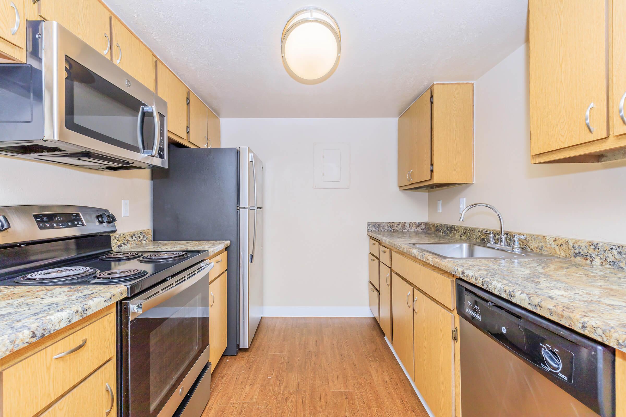 A modern kitchen featuring wooden cabinets, stainless steel appliances including a microwave, oven, refrigerator, and dishwasher. The countertops are made of granite, and the flooring is a light wood finish. The kitchen is well-lit with a ceiling light fixture and has a clean, spacious layout.