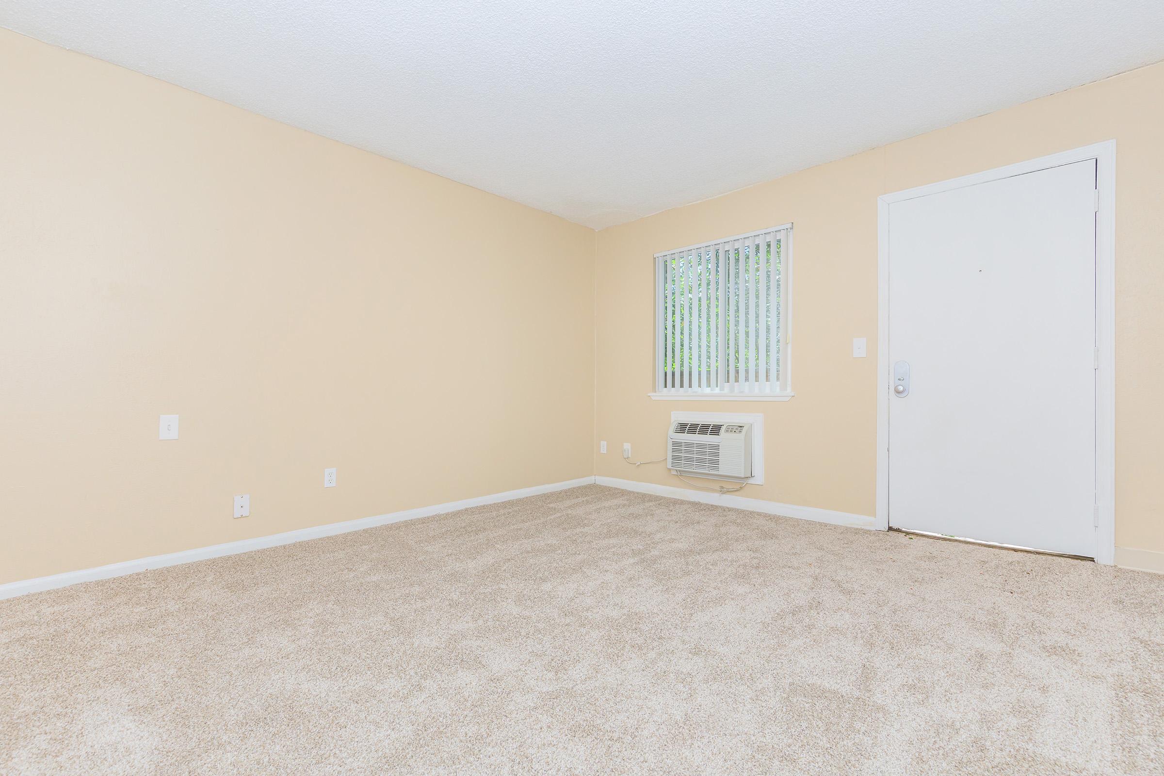 A vacant room with beige walls and light beige carpet. There is a window with blinds on the left side and an air conditioning unit beneath it. The door on the right side is closed, and the room appears bright and clean, ready for furnishing.