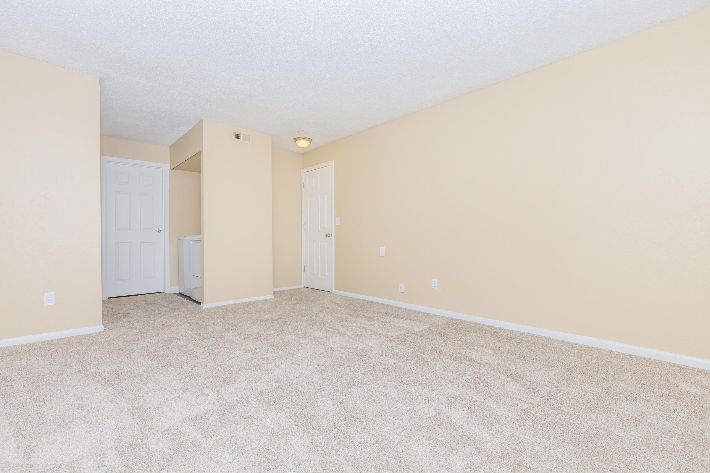 A sparsely furnished room with beige walls and carpet. There is a doorway on the left leading to another room, and a space with a washer or dryer in the corner. The overall atmosphere is bright and open, suitable for various layout options.