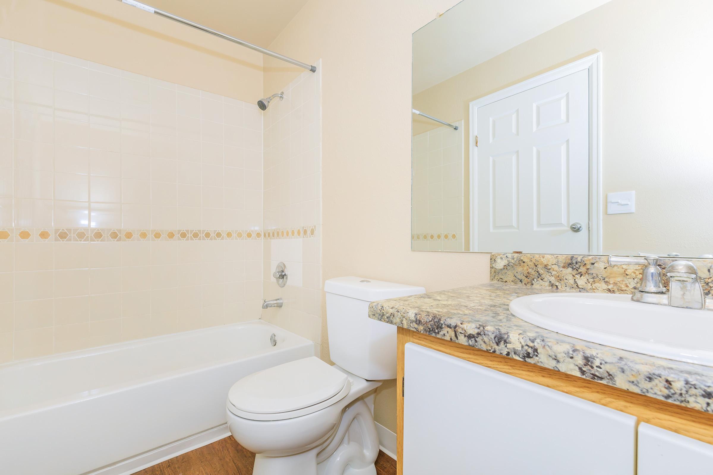 A clean and modern bathroom featuring a bathtub with a shower, a toilet, and a vanity sink with a granite countertop. The walls are painted a light color, and there is a mirror above the sink. A door is visible in the background, and there is a decorative tile border around the tub area.
