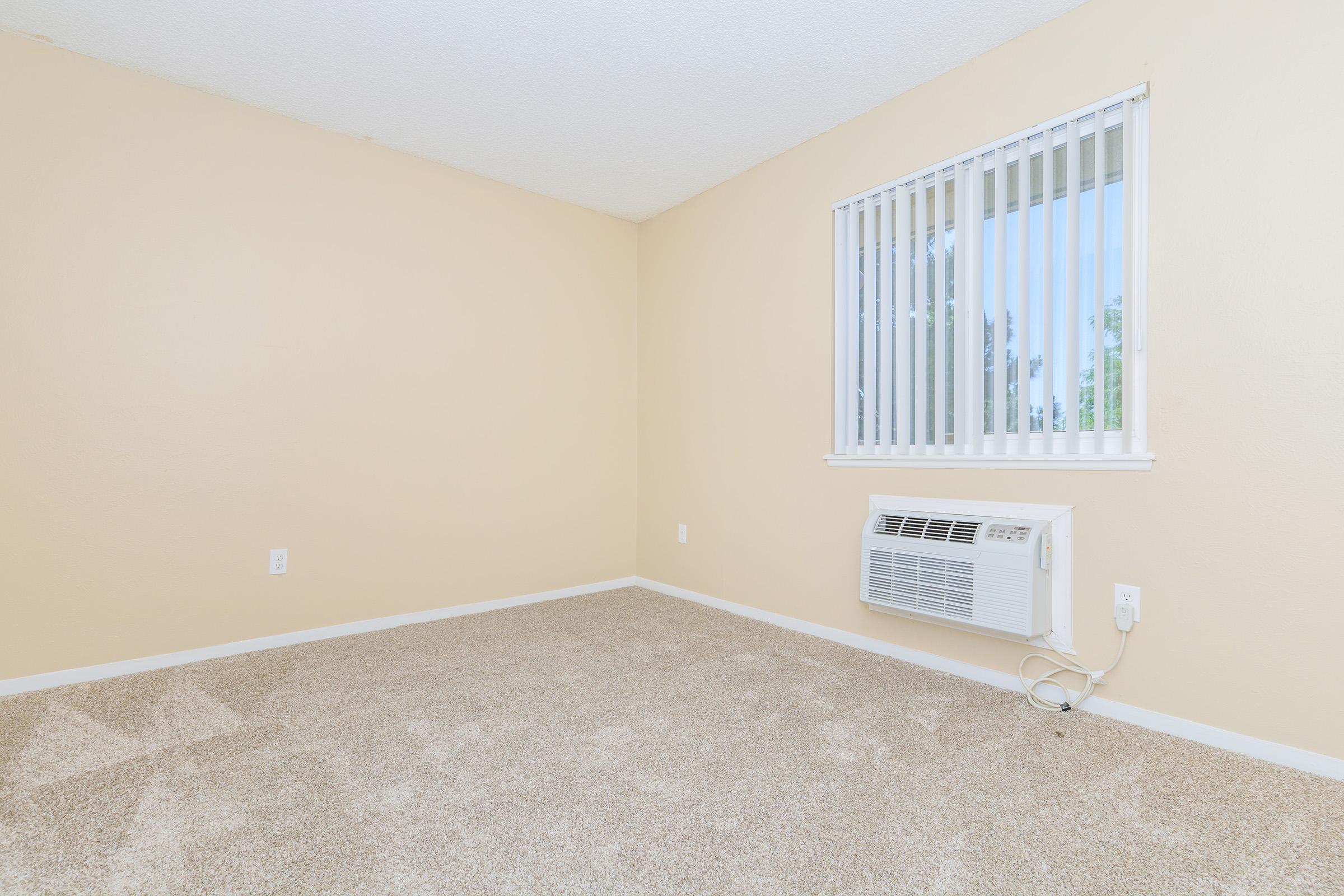 A bright, empty room with beige walls and light-colored carpet. There's a window with vertical blinds allowing natural light, and an air conditioning unit mounted on the wall. The space appears clean and ready for furnishings or decoration.