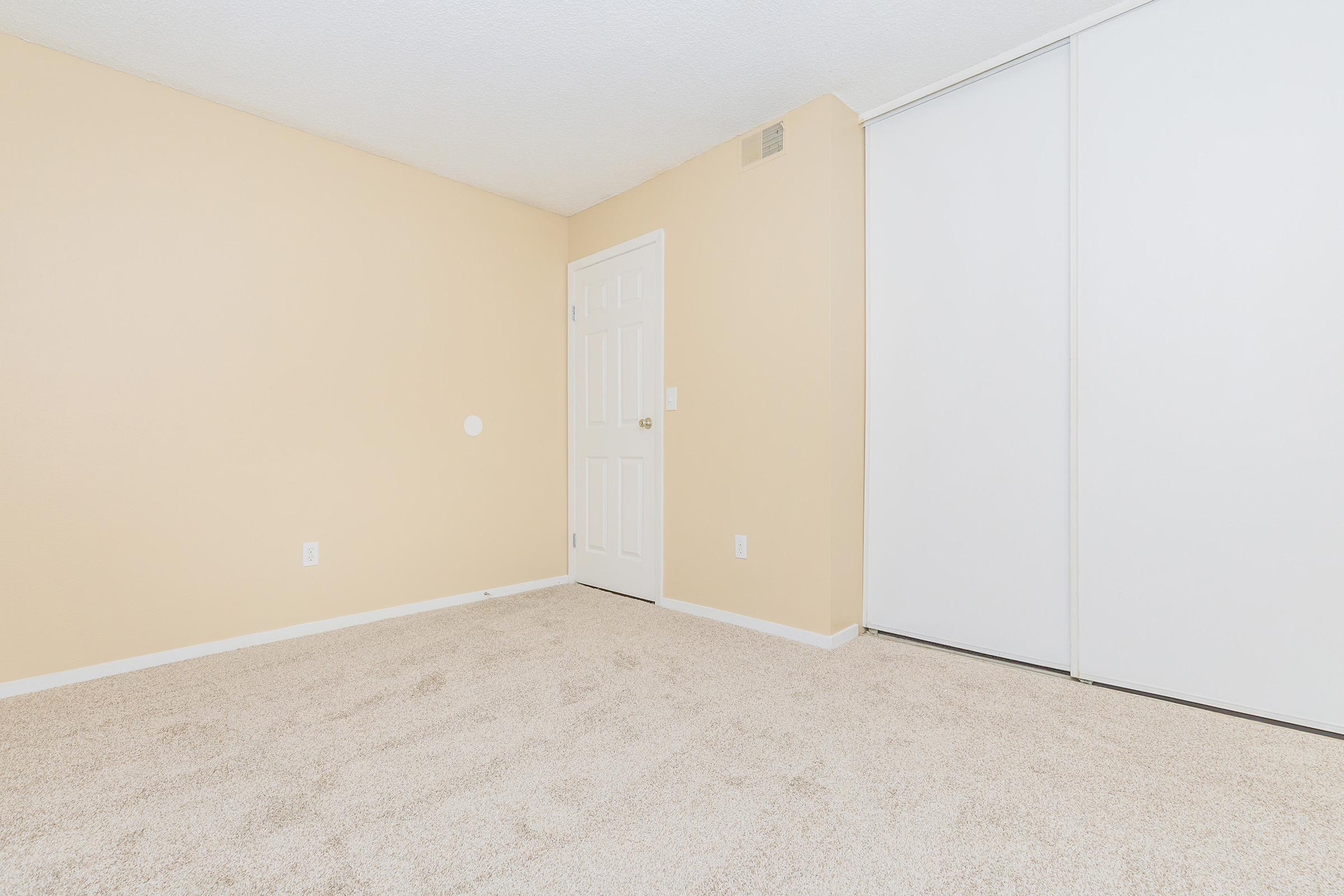 A minimalist, empty room with beige walls and light carpet. The space features a white closet with sliding doors and a closed interior door on the left. Natural light illuminates the room, creating a clean, inviting atmosphere suitable for various uses.