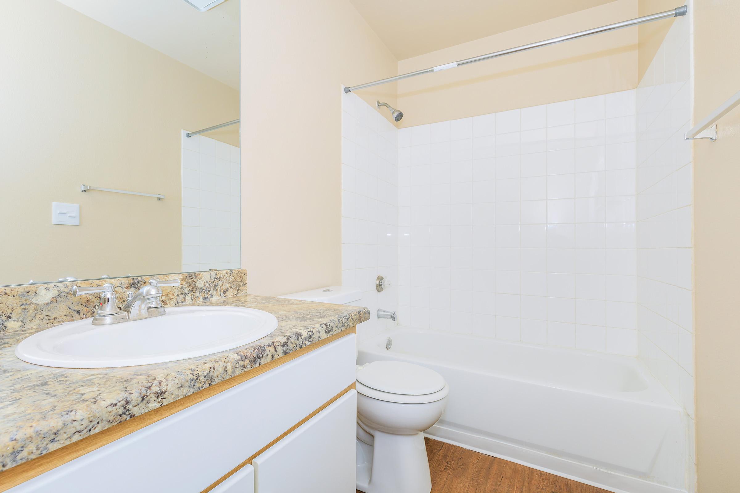 A clean, modern bathroom featuring a white bathtub and shower combination, a single sink with a granite countertop, a toilet, and light-colored walls. A large mirror reflects the space, creating an open and bright atmosphere. The flooring is a light wood grain.