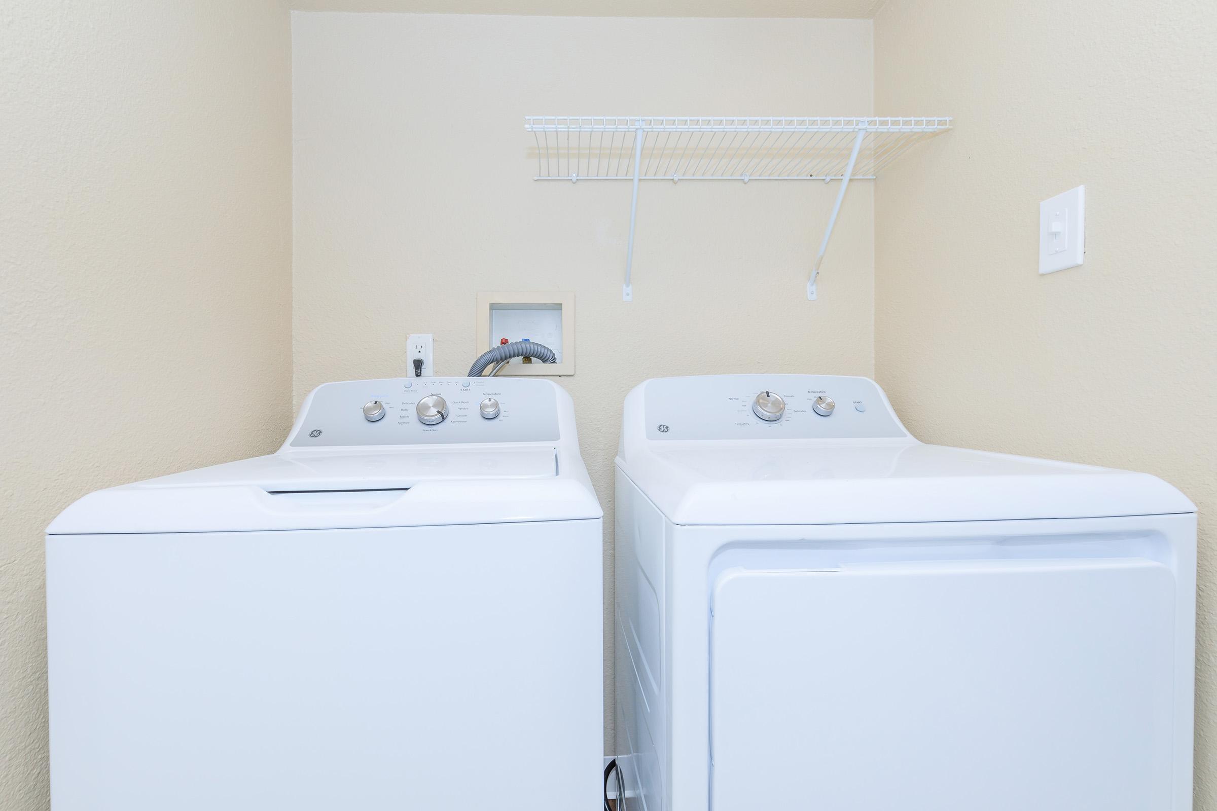 A clean laundry room featuring a white washing machine and a white dryer side by side. Above them, there is a metal shelf for storage, and the wall is painted a light beige color. The room is well-lit, emphasizing the modern appliances.