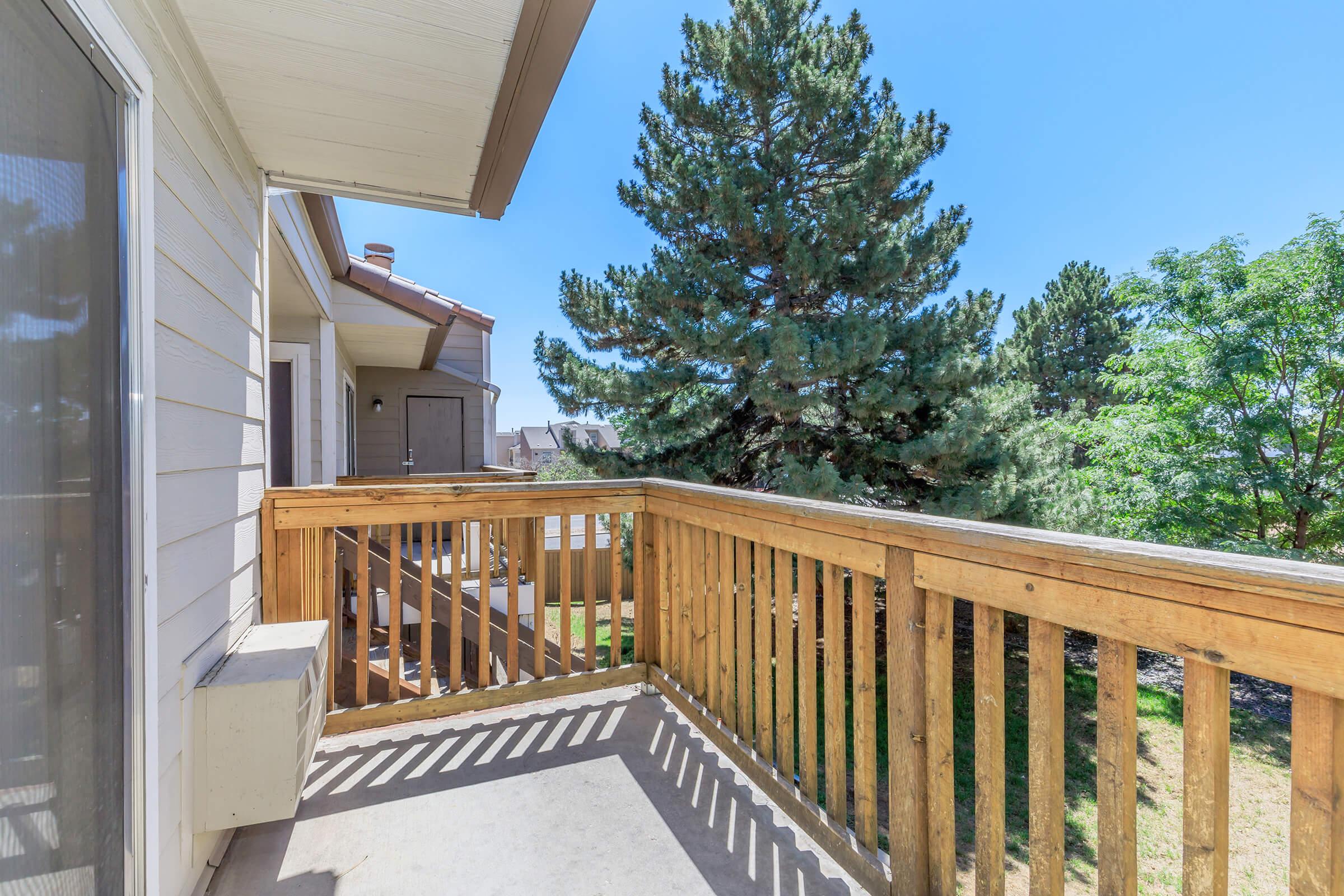 A wooden balcony with a railing, overlooking a green tree and surrounding landscape. The clear blue sky is visible above, and the exterior of a building can be seen in the background. The balcony area is well-lit by sunlight.