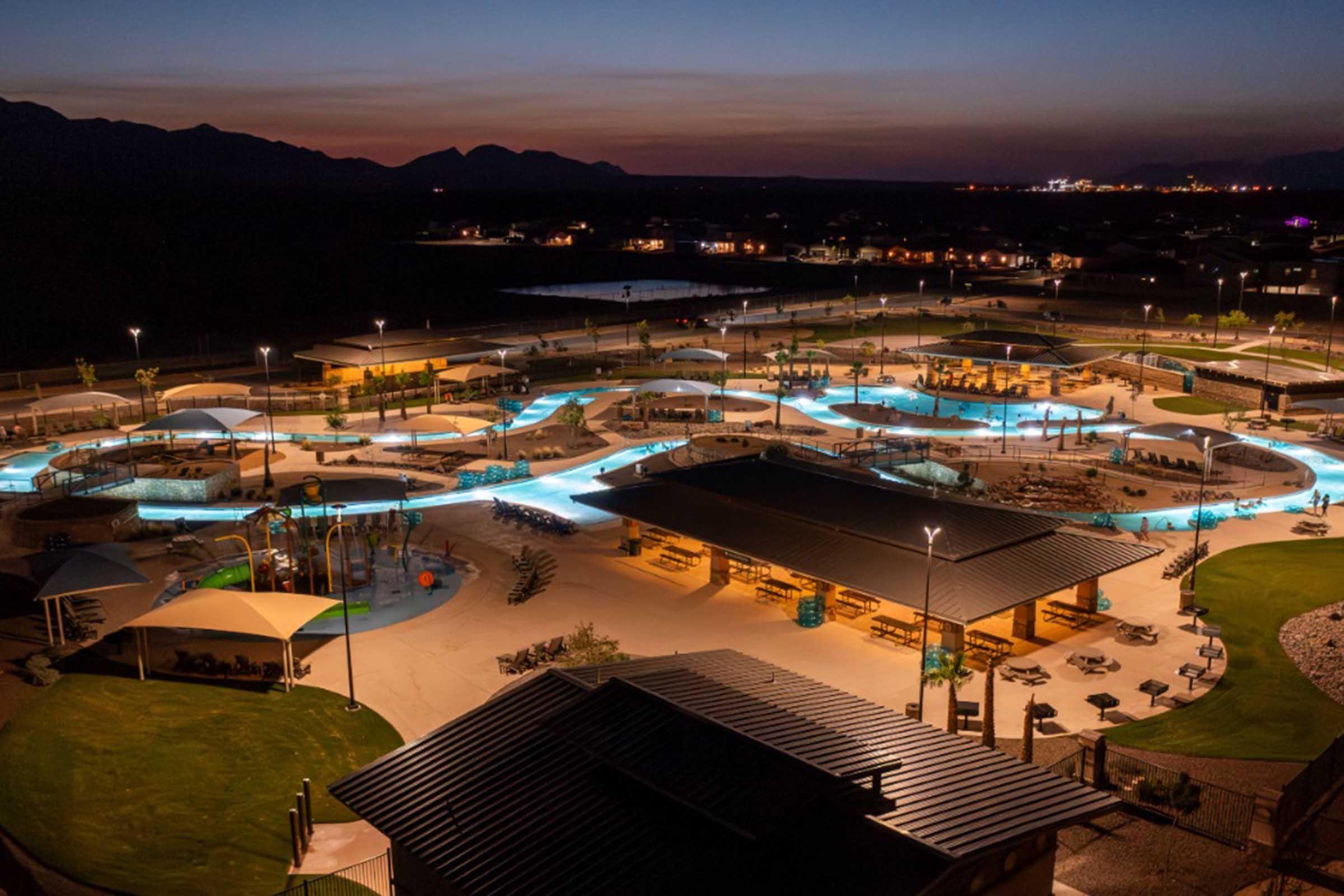 Aerial view of a modern water park illuminated at night, featuring winding water slides, lounging areas, and palm trees. Surrounding the park are illuminated buildings and a scenic landscape with mountains in the background, creating a vibrant and inviting atmosphere.