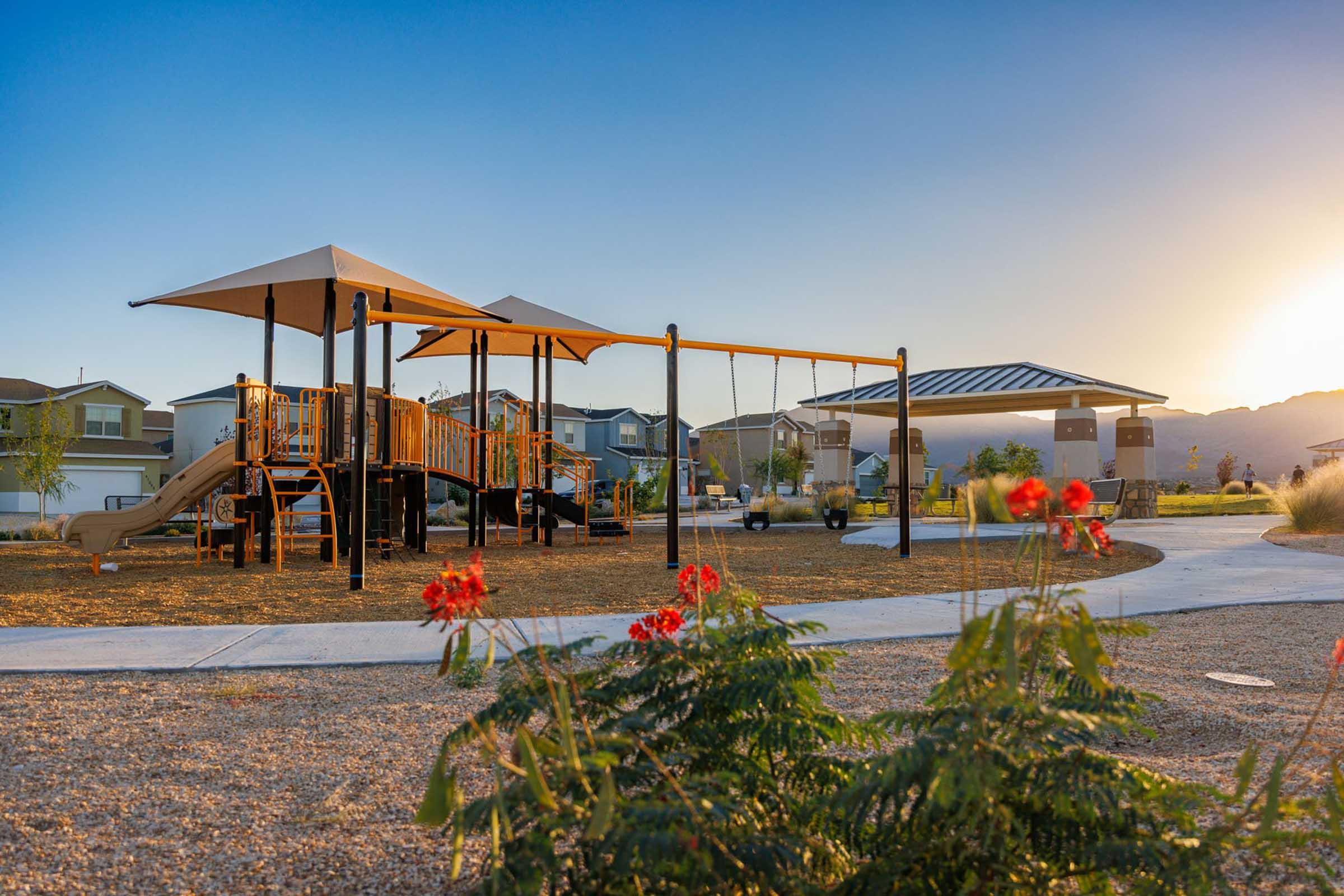 A playground featuring a wooden play structure with slides and swings, set against a backdrop of residential homes. In the foreground, vibrant red flowers grow along a winding path, illuminated by the warm glow of the sunset in the distance.
