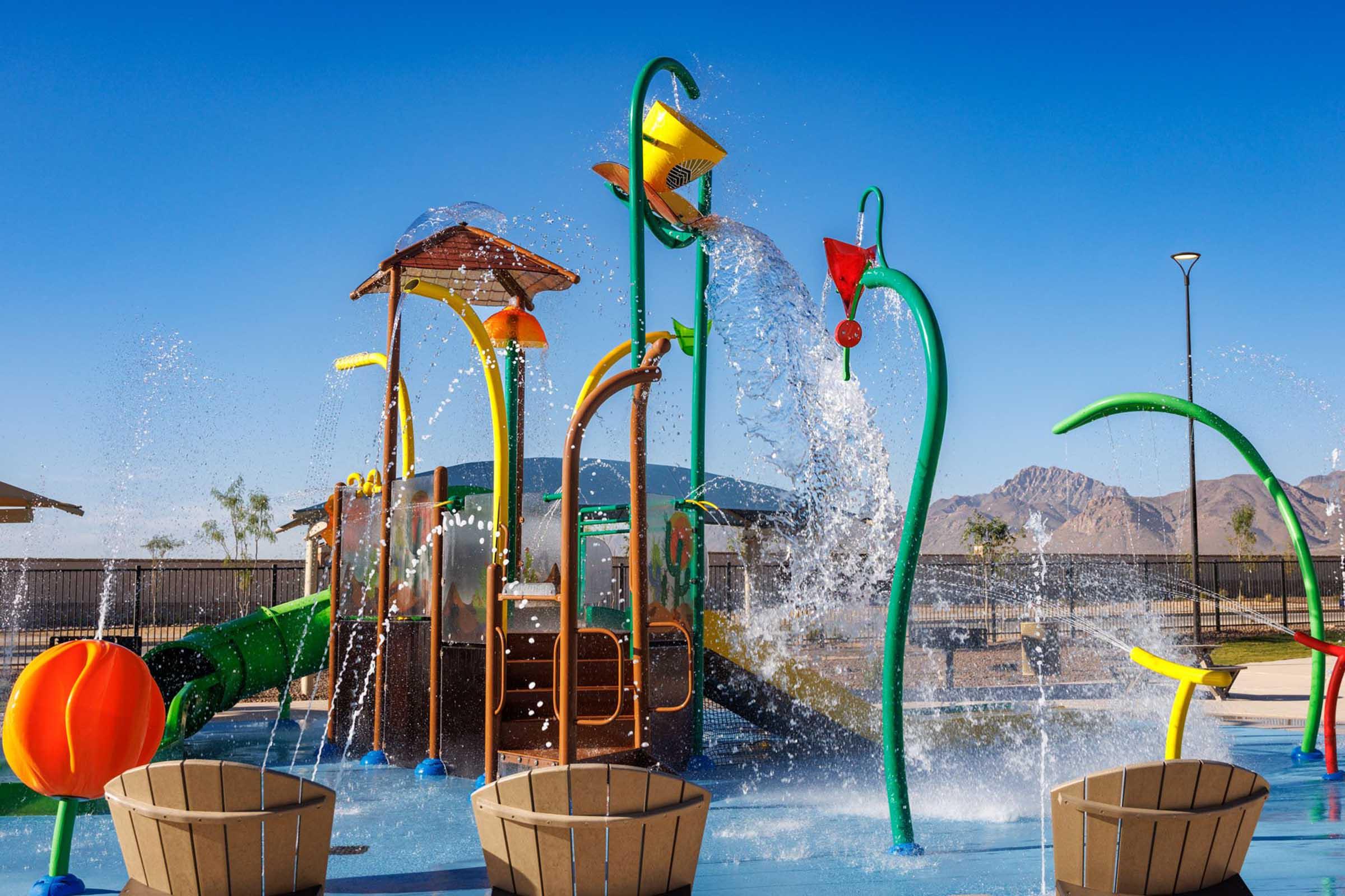 Colorful water play structure at a park featuring slides, splash features, and water jets. Surrounding area includes benches and a clear blue sky, with mountains in the background. Ideal for children's recreational activities on a sunny day.